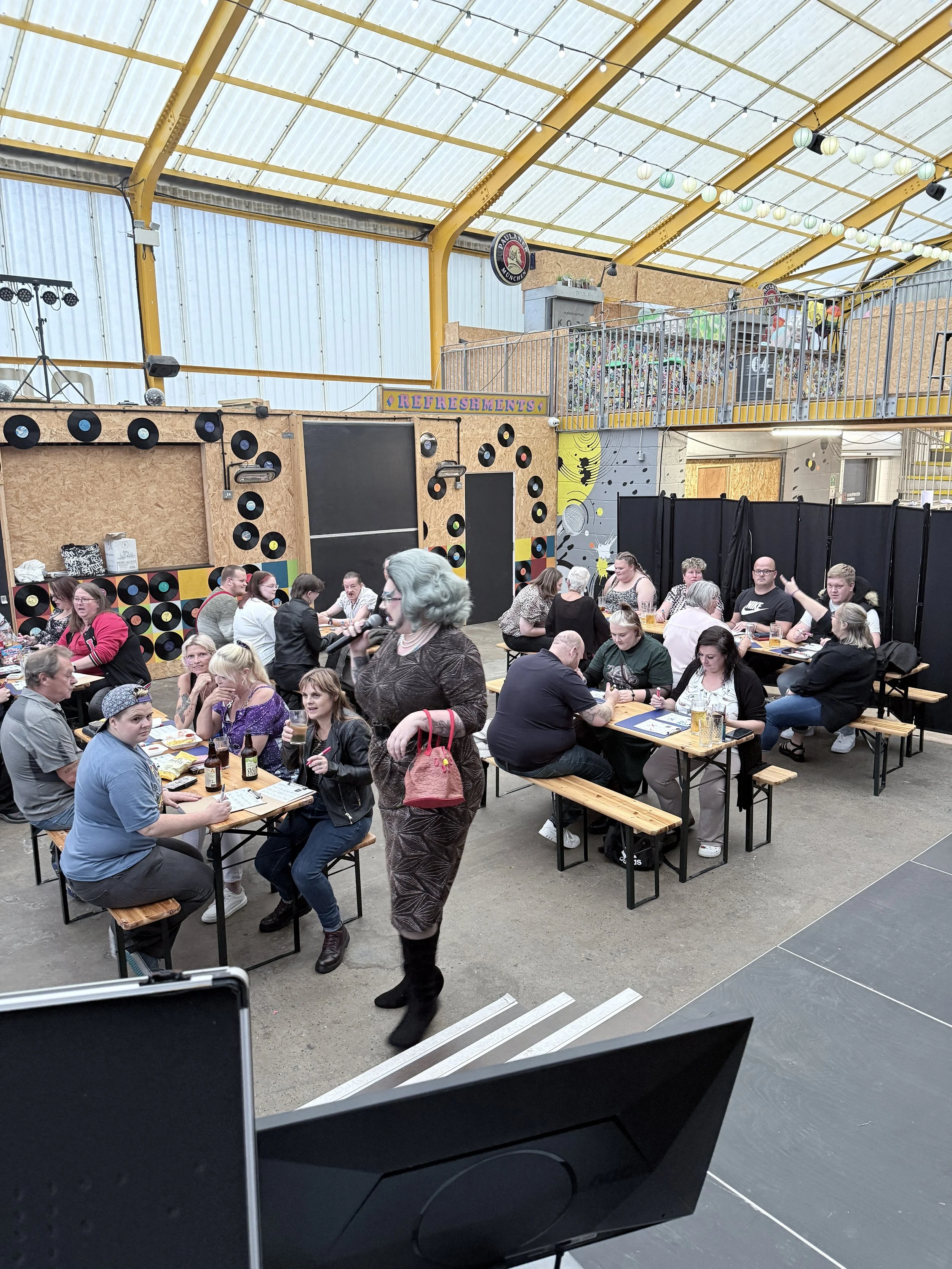 Indoor event space filled with people sitting at tables, a woman holding a microphone, decorated with vinyl records and vintage decor, with a high roof and string lights.