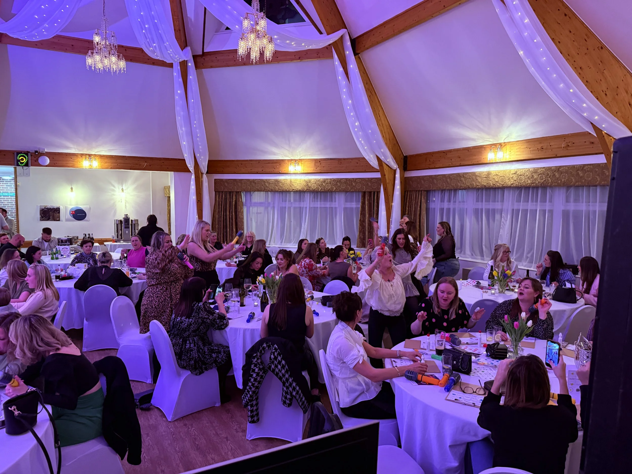 An indoor event space decorated with white drapes and chandeliers, filled with round tables and people, some taking photos or holding microphones, likely at a celebration or gathering.