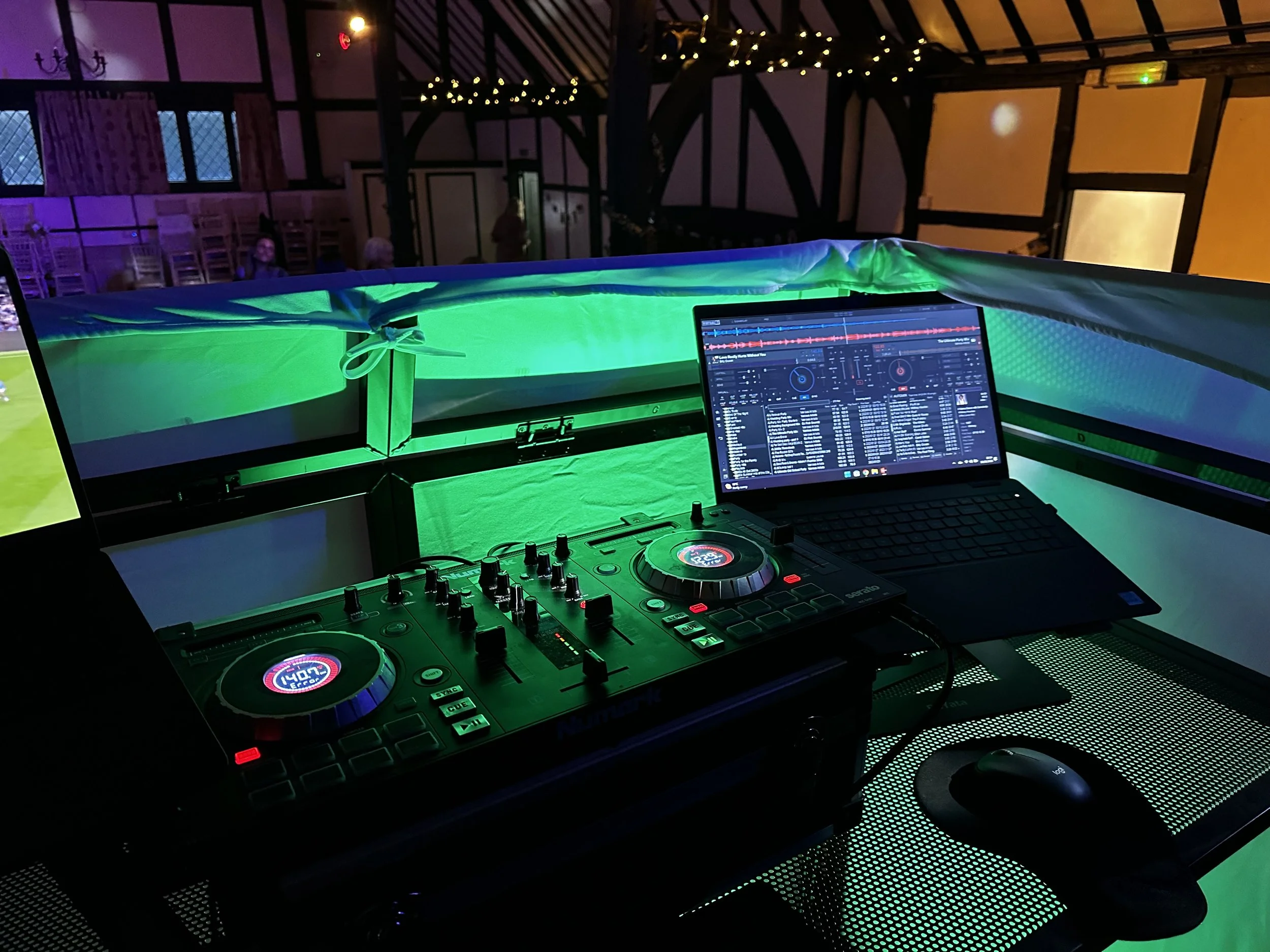 DJ setup with turntables, a laptop with DJ software, and a green-lit booth in a dimly lit venue with decorative lighting on the ceiling.