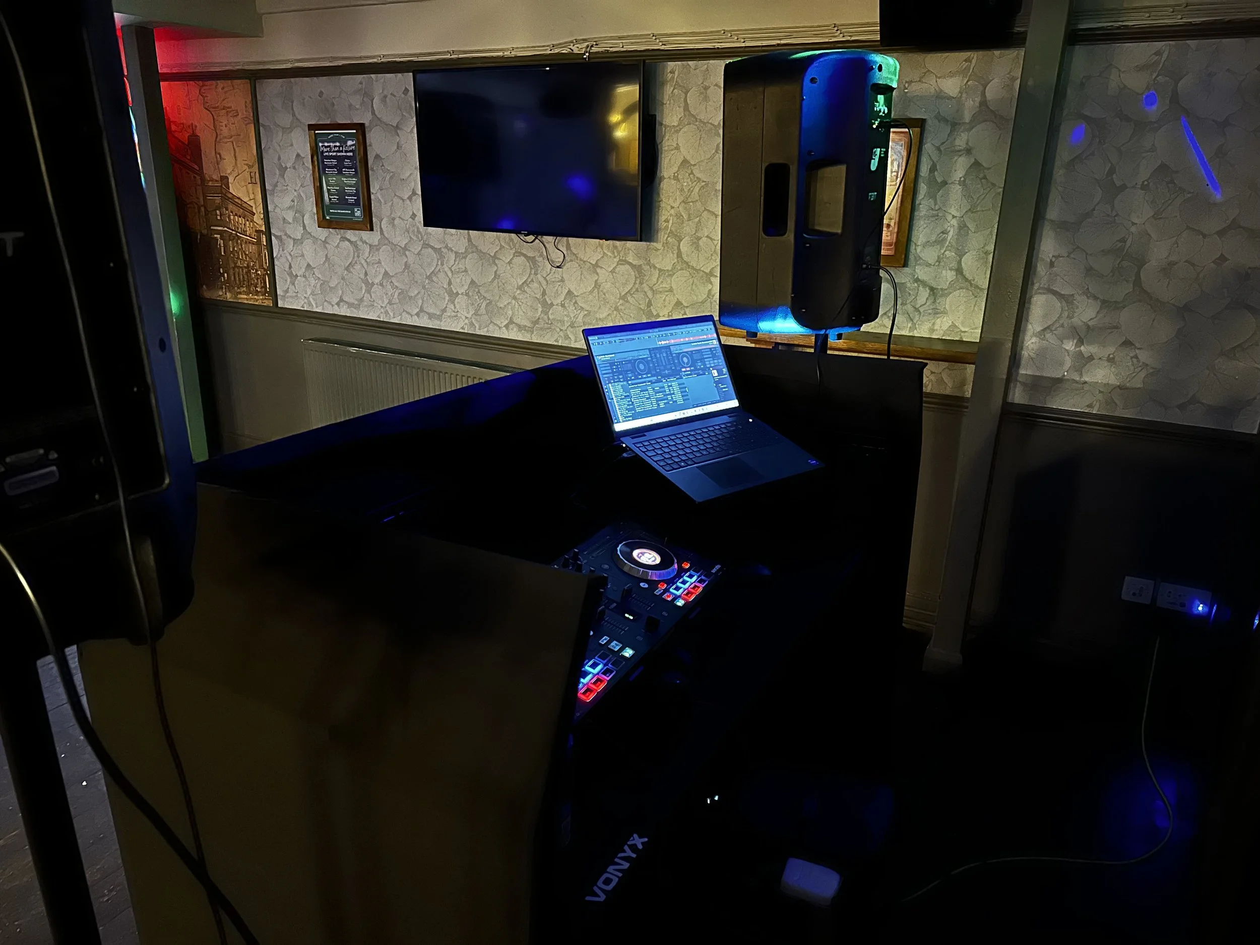 A DJ setup with a laptop, DJ controller, and speakers in a dimly lit room, with a wall-mounted TV and framed pictures in the background.
