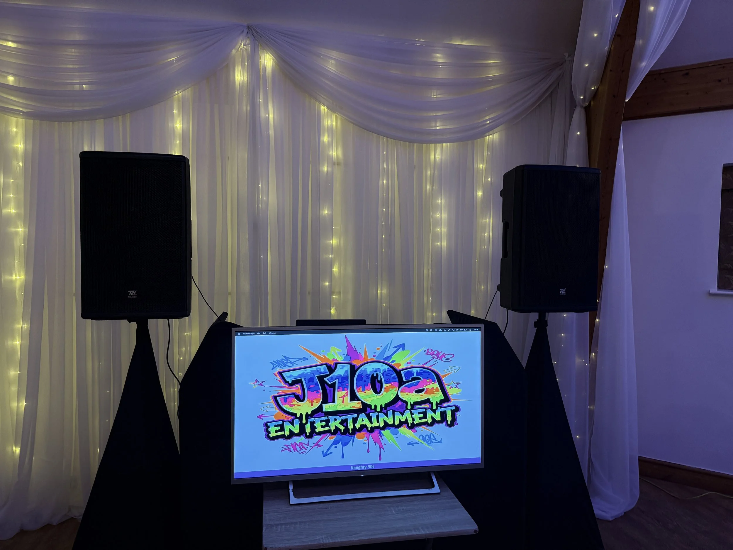 A DJ setup at an event with a backdrop of white curtains decorated with yellow fairy lights. There are two large black speakers and a computer monitor displaying a colorful logo that reads '90s Entertainment' with a graffiti-style design.