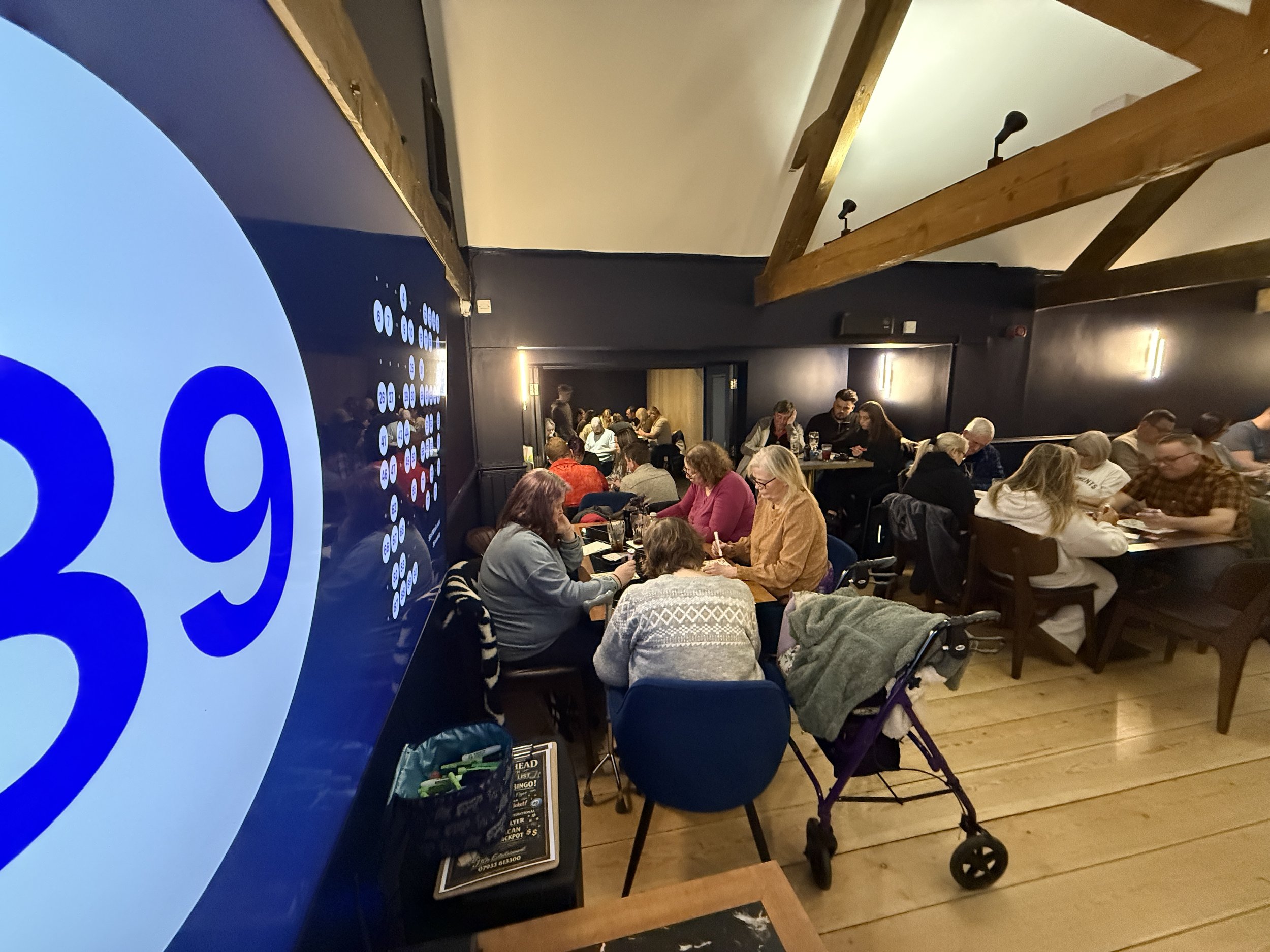 People sitting and standing in a crowded indoor space with tables, some using mobile phones. A large digital screen displaying a Bingo game is partially visible on the left side.