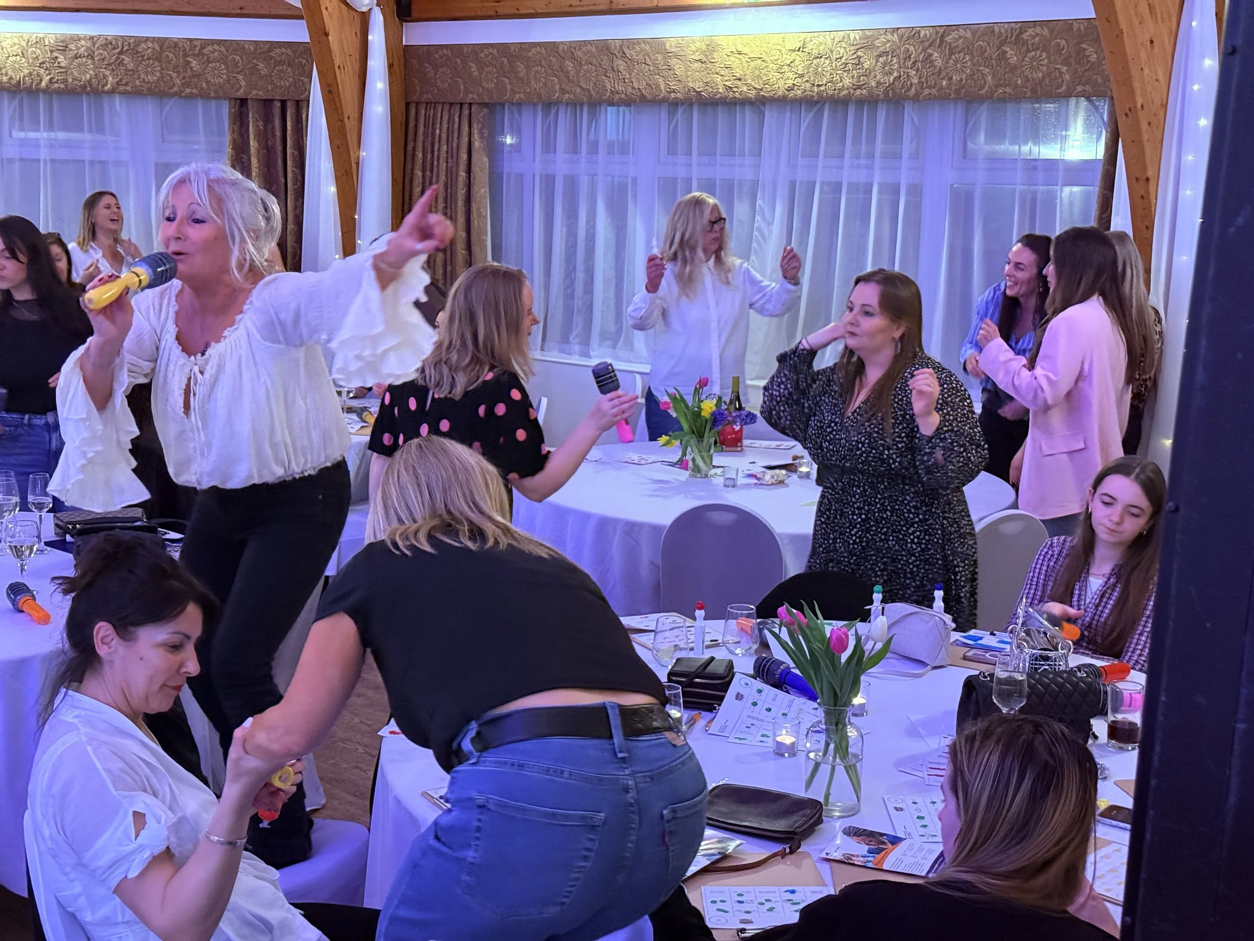 A woman with curly gray hair singing into a yellow microphone, surrounded by women at a dinner party, some listening, some dancing, in a decorated indoor setting with large windows and flowers on the table.