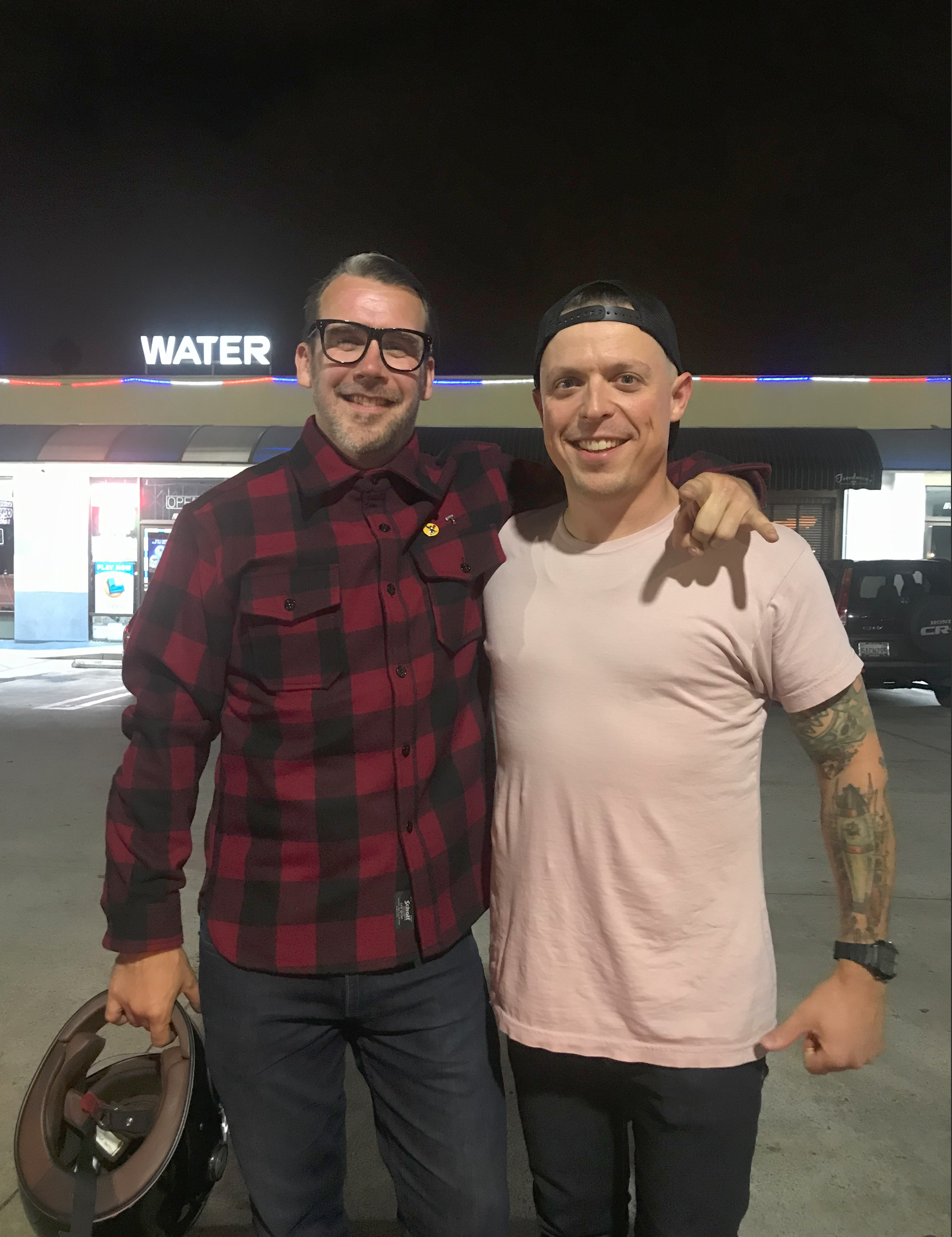 Two men standing side by side outside at night smiling, with one wearing a black backward cap, a pink t-shirt, and visible tattoos on his right arm, and the other wearing glasses, a red and black plaid shirt, with a person's backpack and a brown helmet hanging on his left arm. There is a lit sign that says 'WATER' in the background.