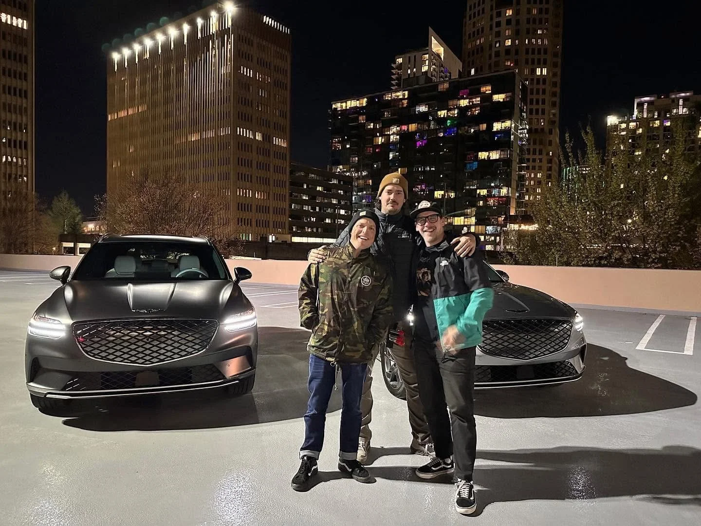 Three young men stand arm-in-arm in a parking lot at night, with two modern black cars behind them and a city skyline with tall illuminated buildings in the background on a video production shoot