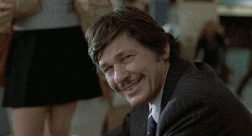 A man with brown hair and a mustache, smiling at the camera, wearing a dark suit. There is a blurred background with people and furniture.