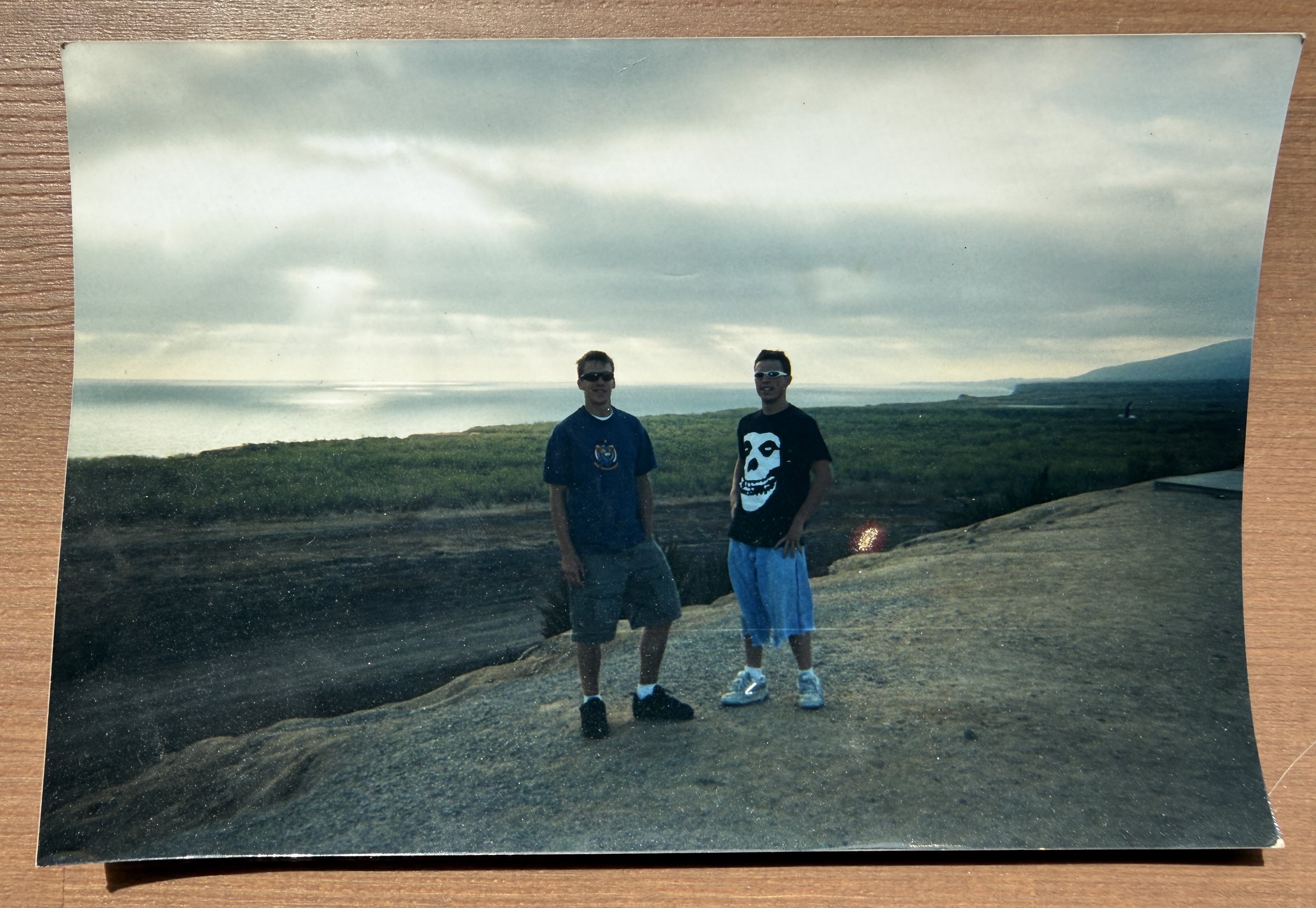Two young men standing outdoors on a rocky area by a body of water, with green grass and hills in the background, under a cloudy sky.