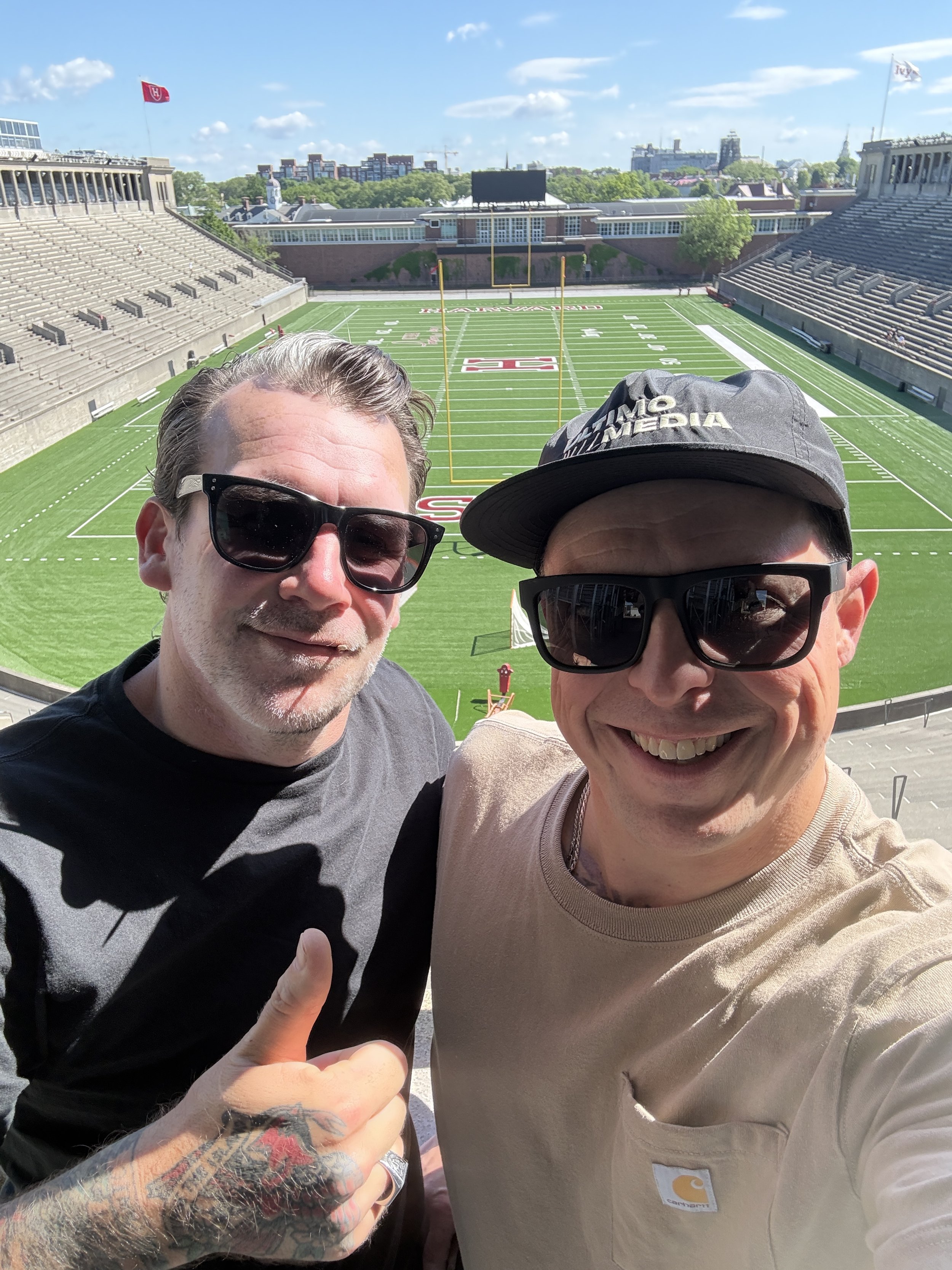 Two smiling men are taking a selfie inside a football stadium. The field and empty stands are visible behind them under a blue sky with some clouds.