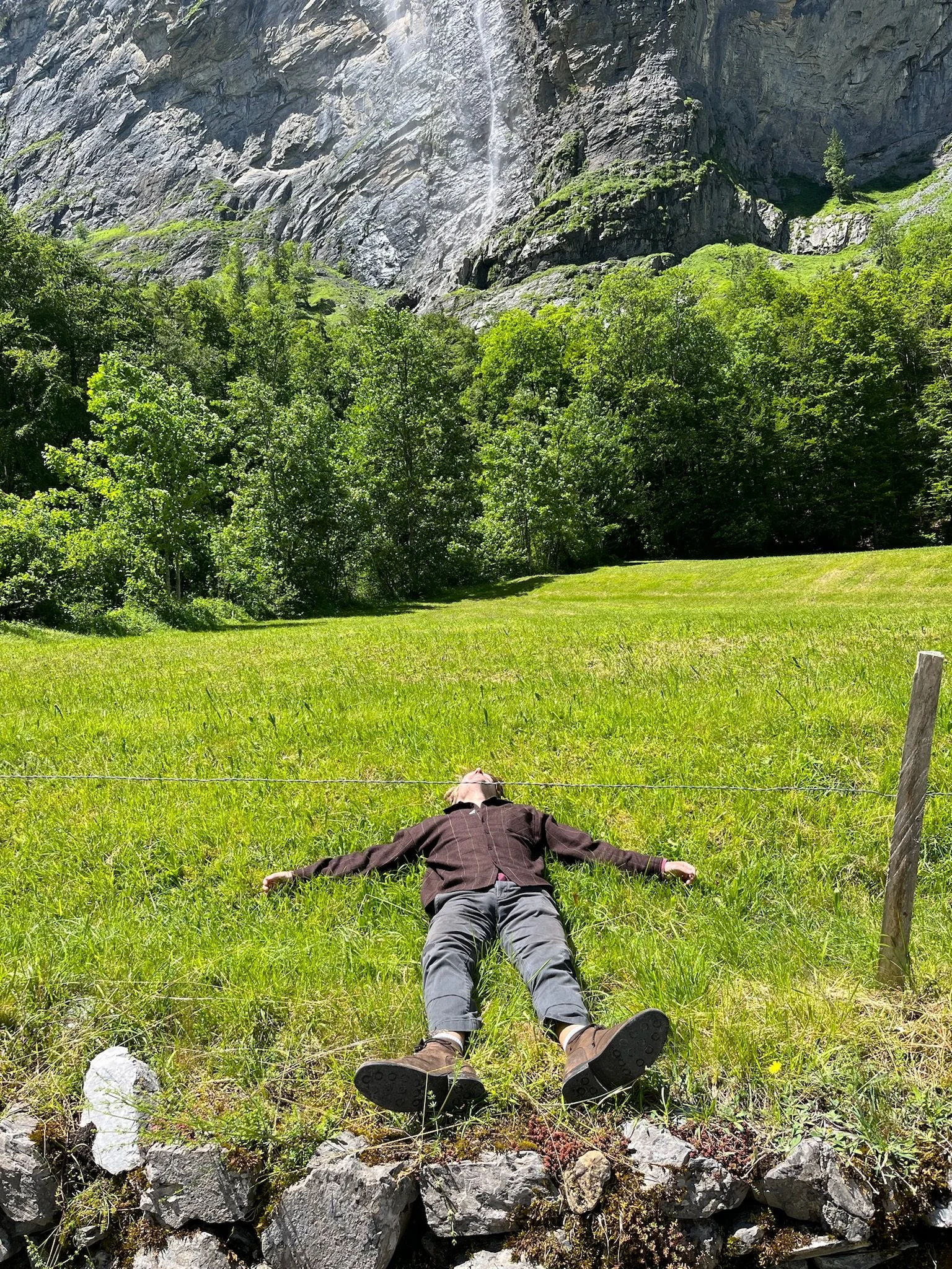 A person laying on their back on a grassy field near rocks, facing upward with arms outstretched, in front of green trees, with mountain cliffs and thin waterfall in the background.