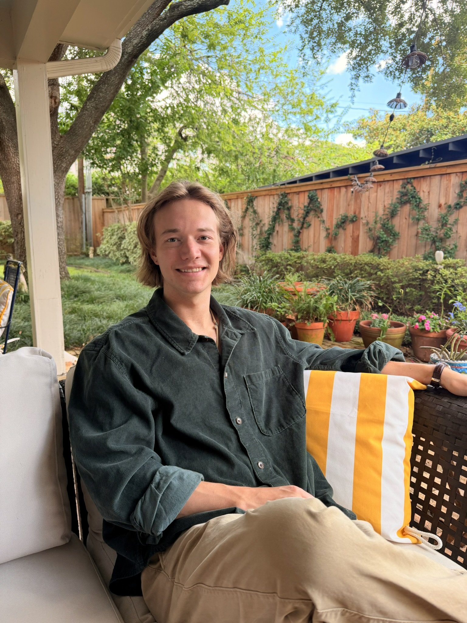 A young man with shoulder-length brown hair sitting outdoors on a patio or porch, smiling at the camera. He is wearing a dark green button-up shirt and beige pants. In the background, there is a wooden fence, potted plants, and trees with green leaves. The sky is partly cloudy with some blue visible. There are string lights hanging above, and a striped yellow and white towel is draped over the armrest of the chair.