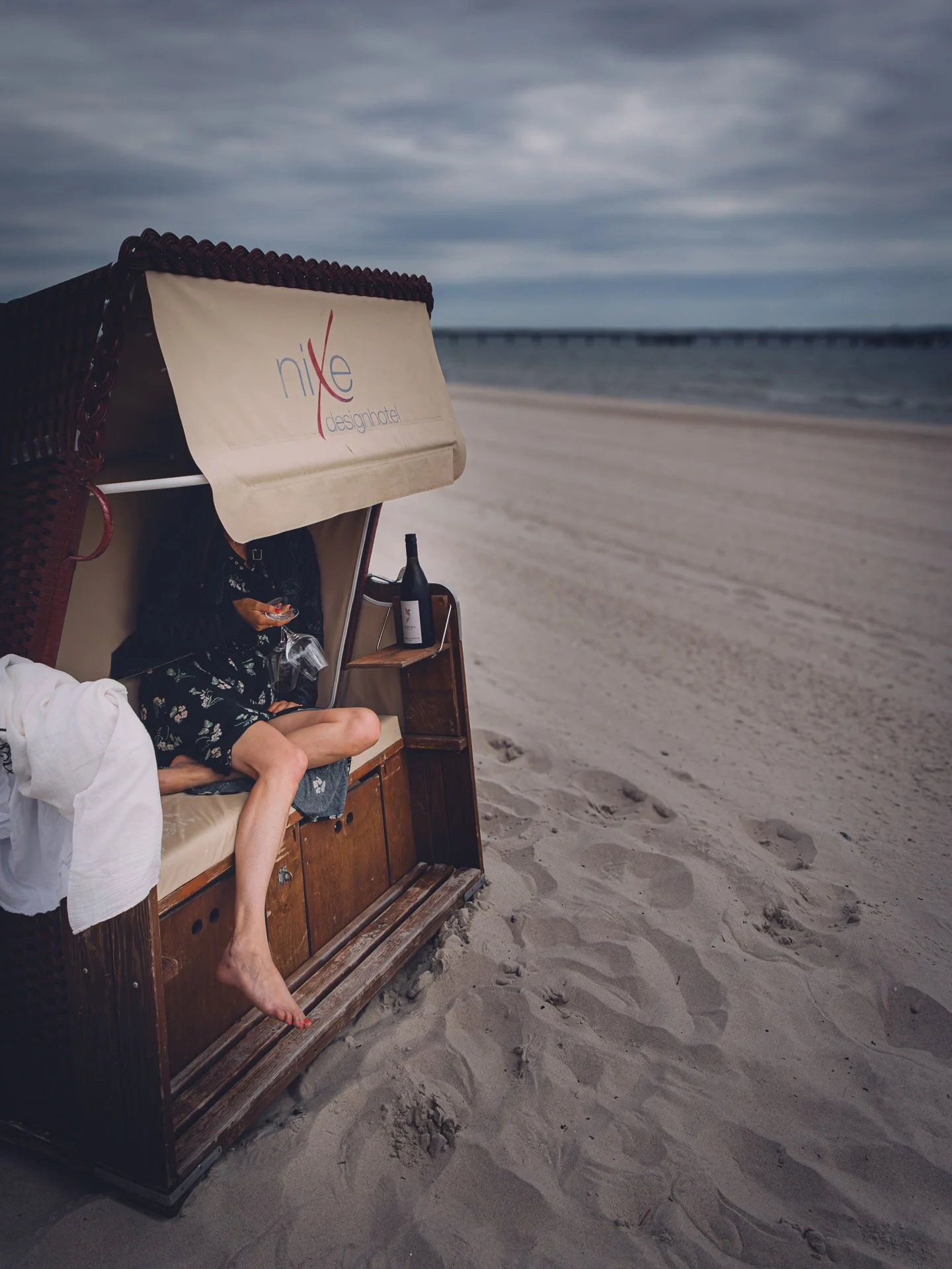 Beachside cabana with a woman sitting inside holding a glass, with a bottle of wine on a small table nearby; cloudy sky and ocean in the background.