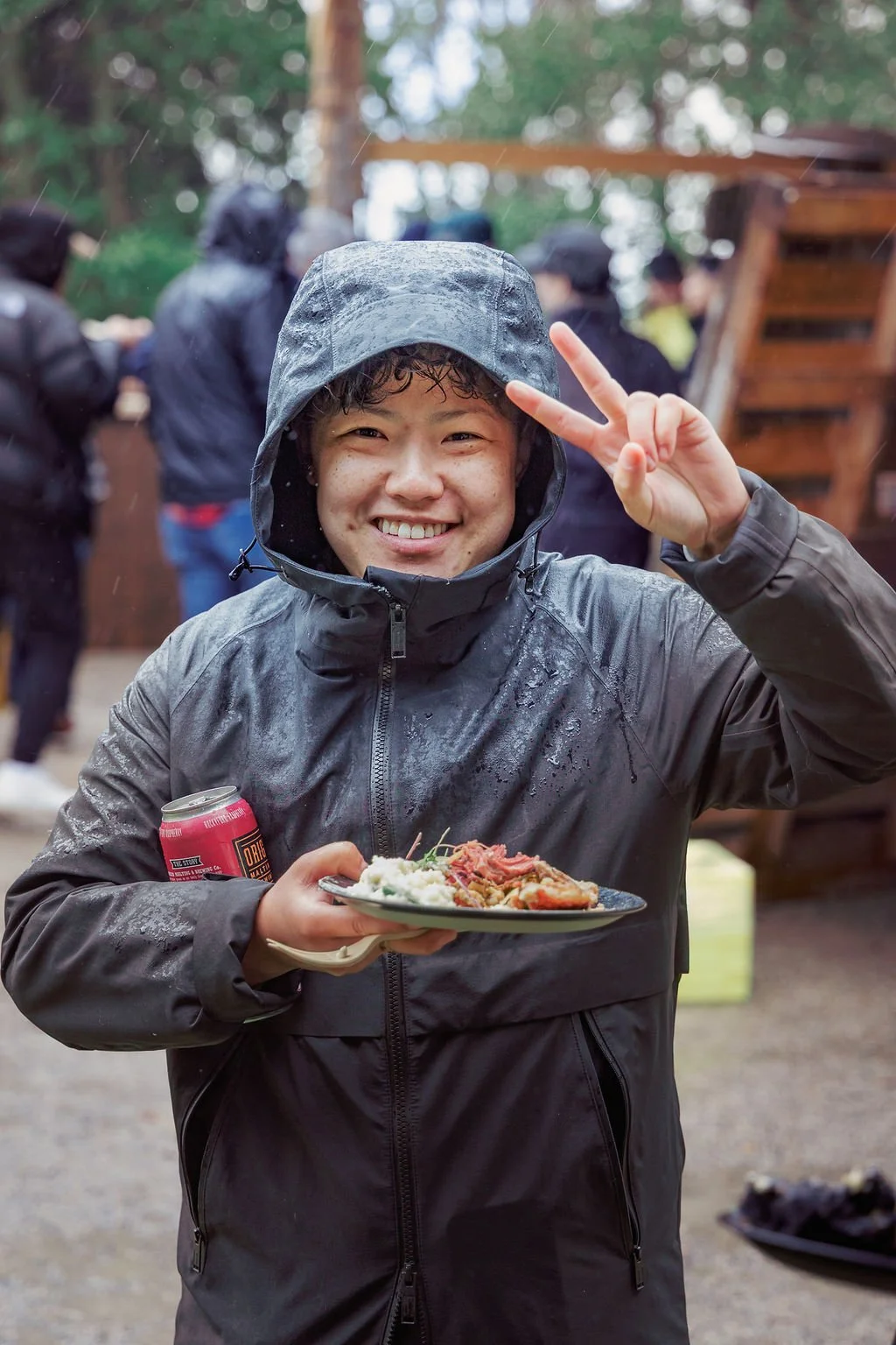A young man in a waterproof jacket and hood, smiling and flashing a peace sign, holding a plate of food and a soda can at an outdoor gathering in rainy weather.