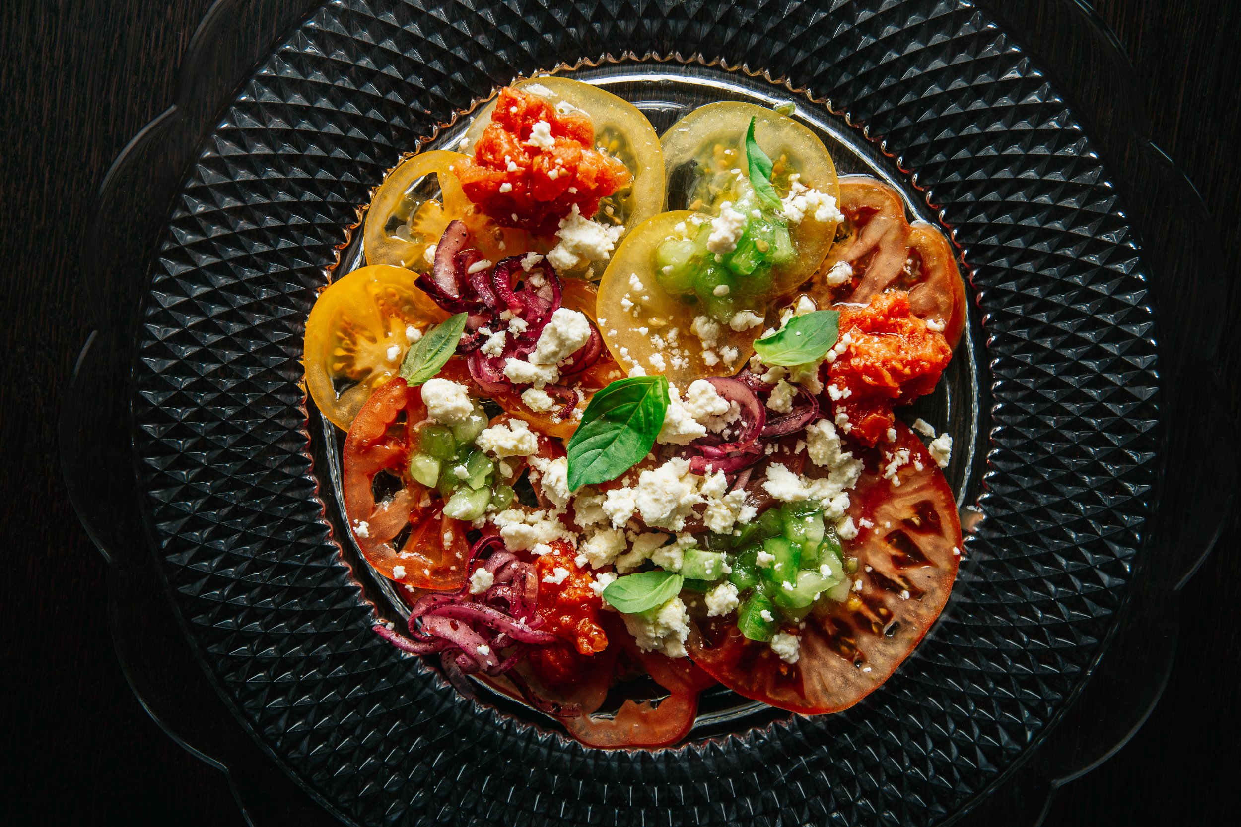 A glass plate with a decorative pattern containing a colorful assortment of sliced heirloom tomatoes topped with crumbled feta cheese and fresh basil leaves, placed on a dark wooden surface.