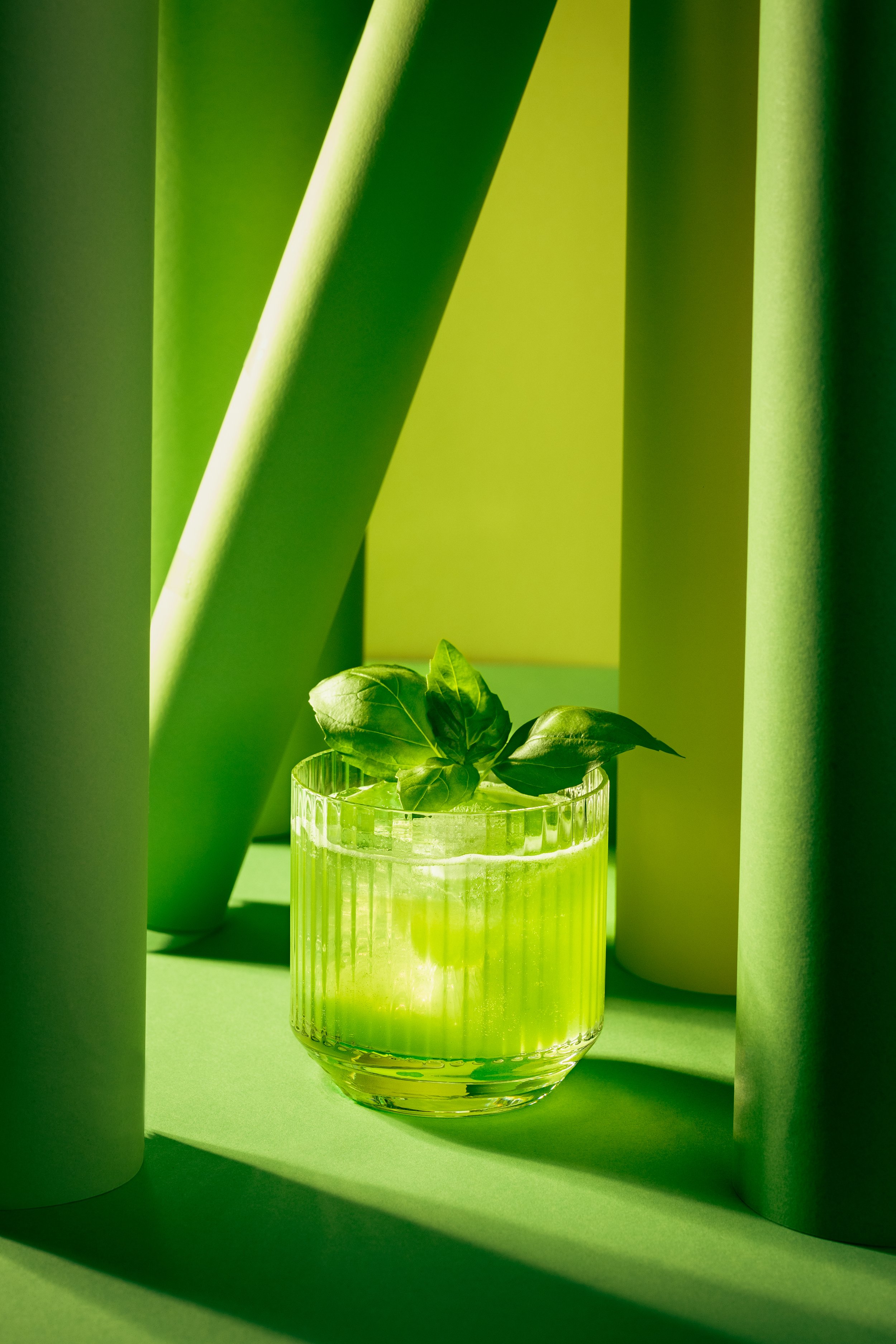 Green-tinted photo of a glass with water and basil leaves, surrounded by tall white cylindrical objects, creating a dappled sunlight effect.