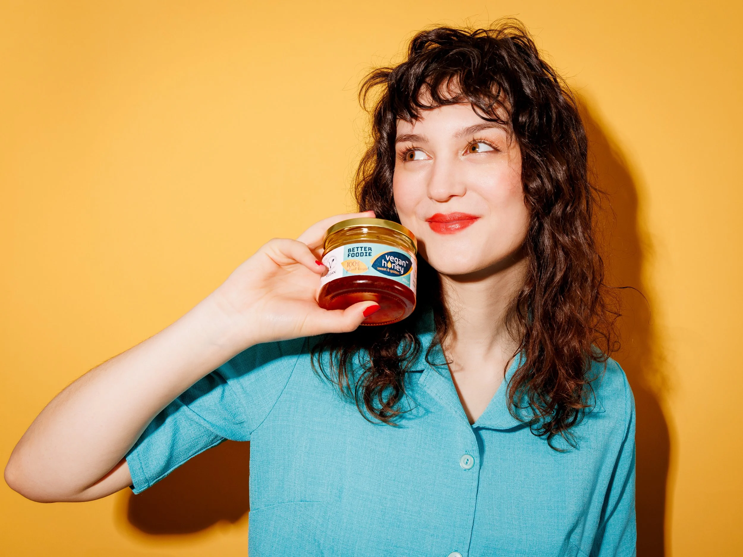 A woman in a turquoise shirt holding a jar of vegan honey near her face against a yellow background.
