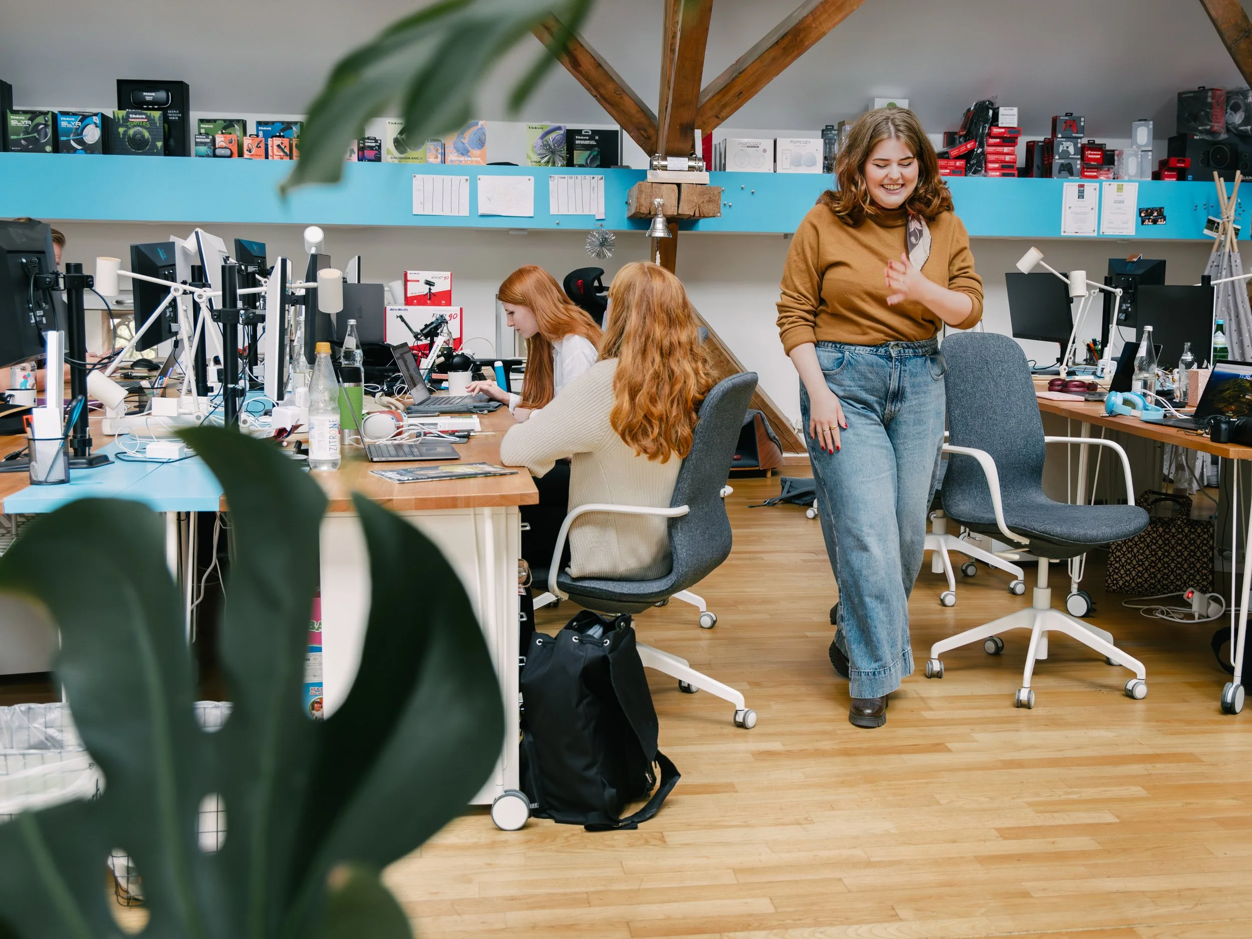 A woman with curly hair in a brown sweater and jeans standing in an office, smiling, with a workspace with computers, laptops, and other office supplies behind her. Two women with red hair working on laptops are seated at desks, and a large plant par