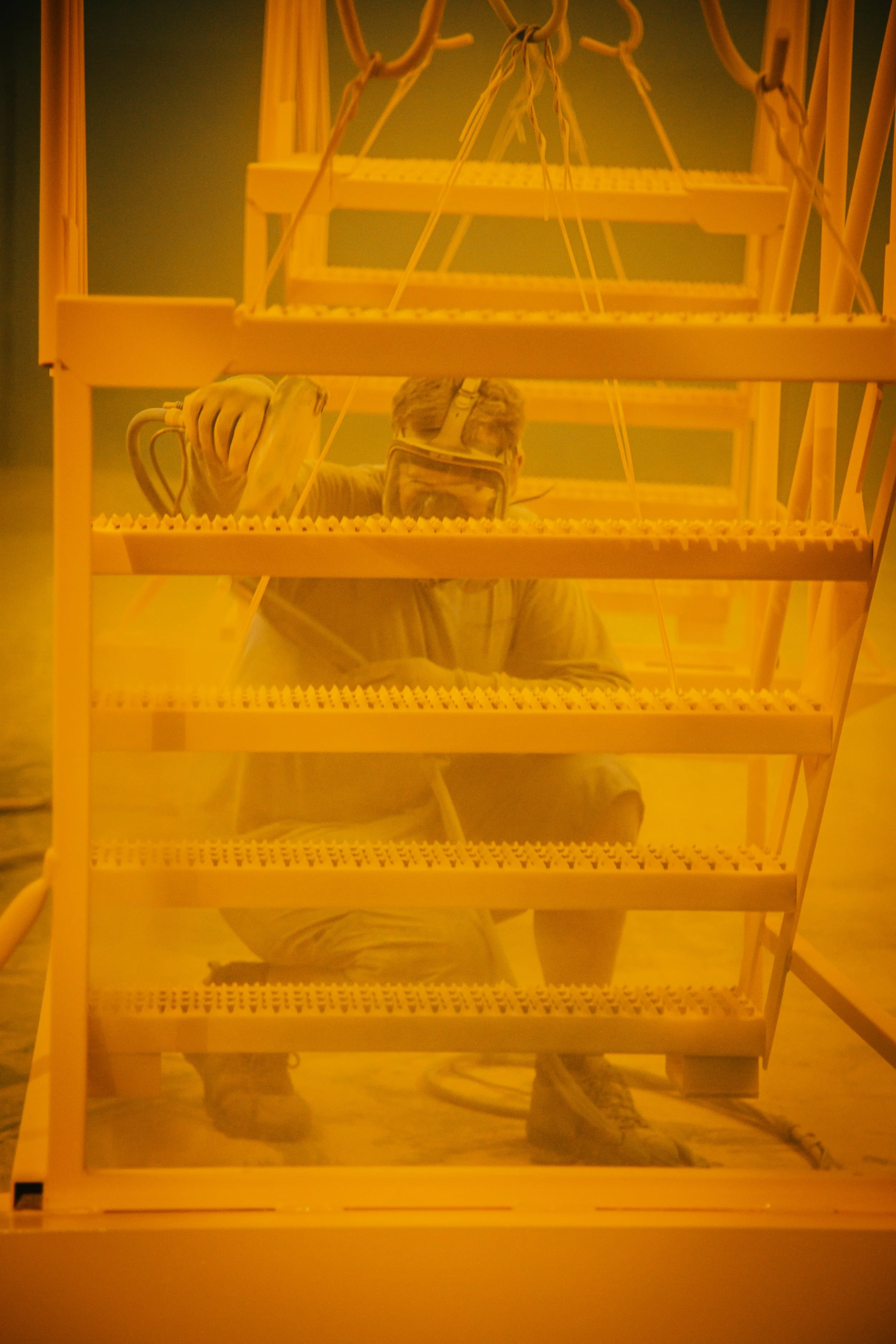 Person wearing a mask and gloves spray-paints a wooden installation with a yellowish hue.