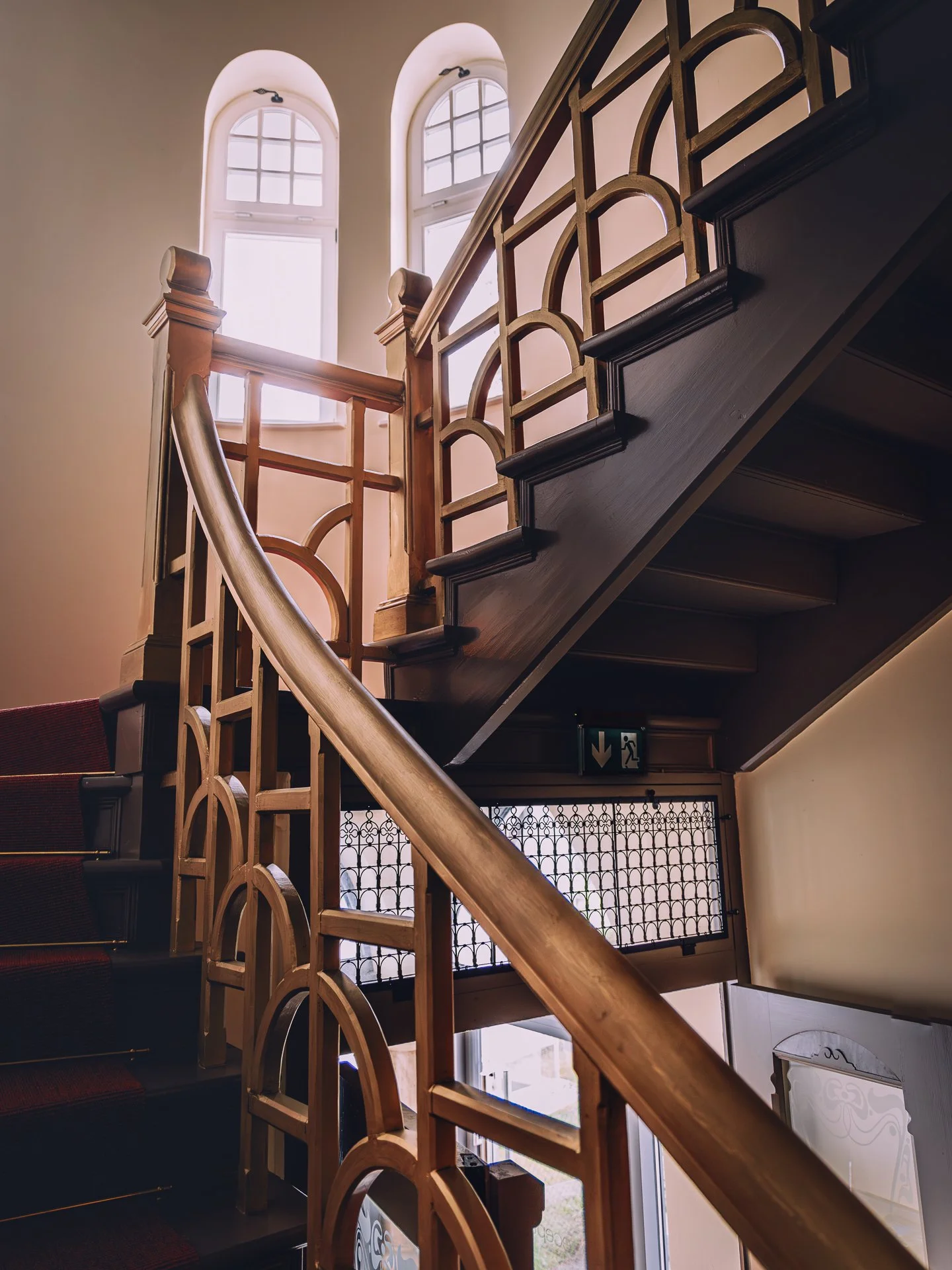Indoor staircase with wooden railing and decorative metal balustrade next to a large arched window.