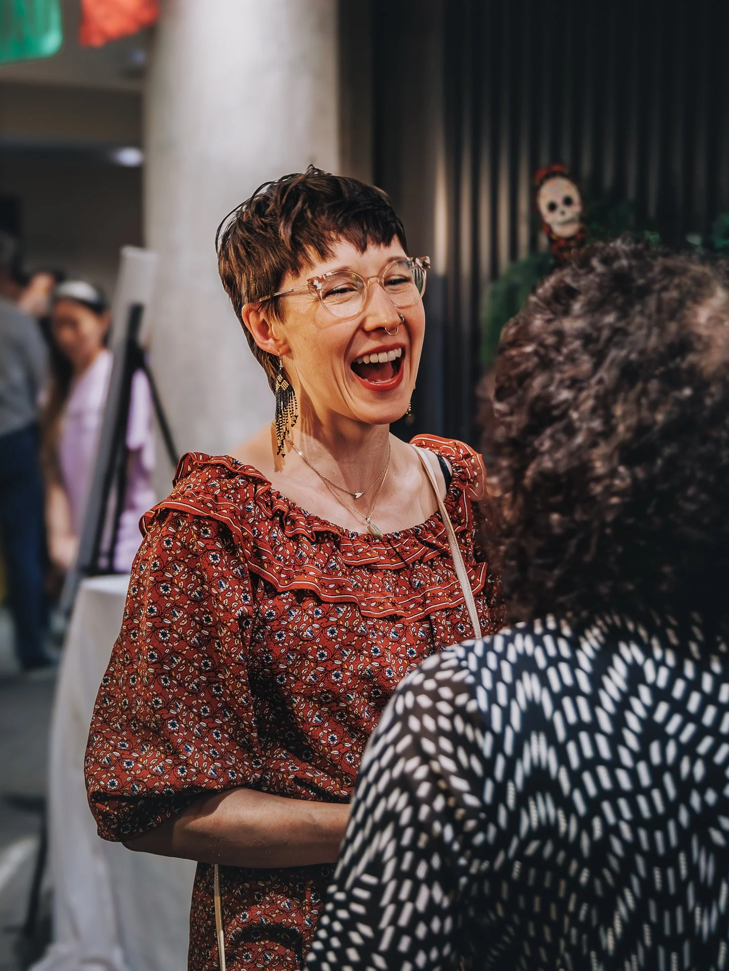 A woman laughing and talking to another person in a social setting. She has short brown hair, glasses, and is wearing a patterned red dress with floral designs, jewelry including earrings and a necklace.