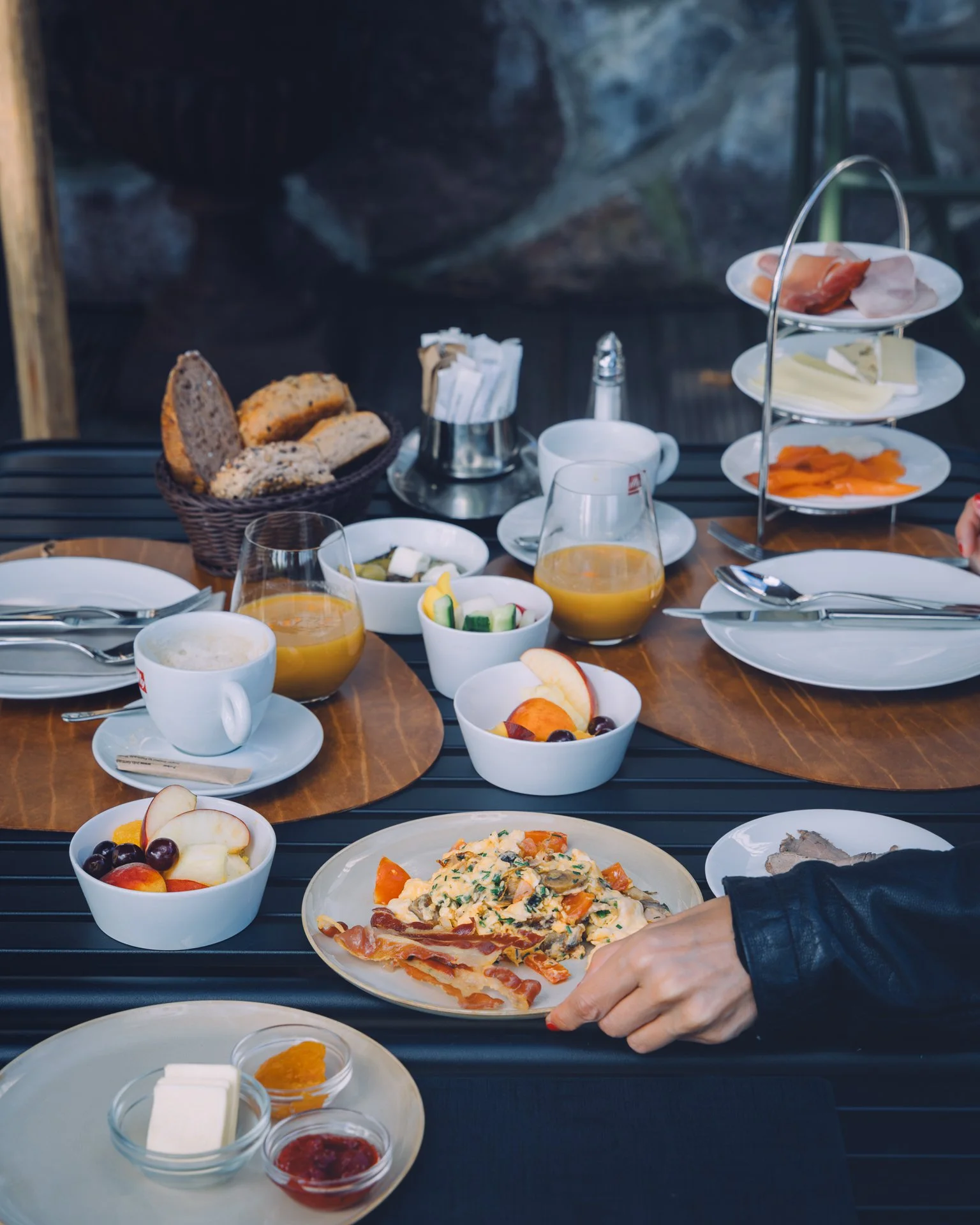 Breakfast spread on a black table includes bowls of fruit, slices of bread, a plate of omelette with vegetables and ham, slices of cheese and meats, small bowls of jam and butter, cups of tea or coffee, a glass of orange juice, and a three-tiered sta