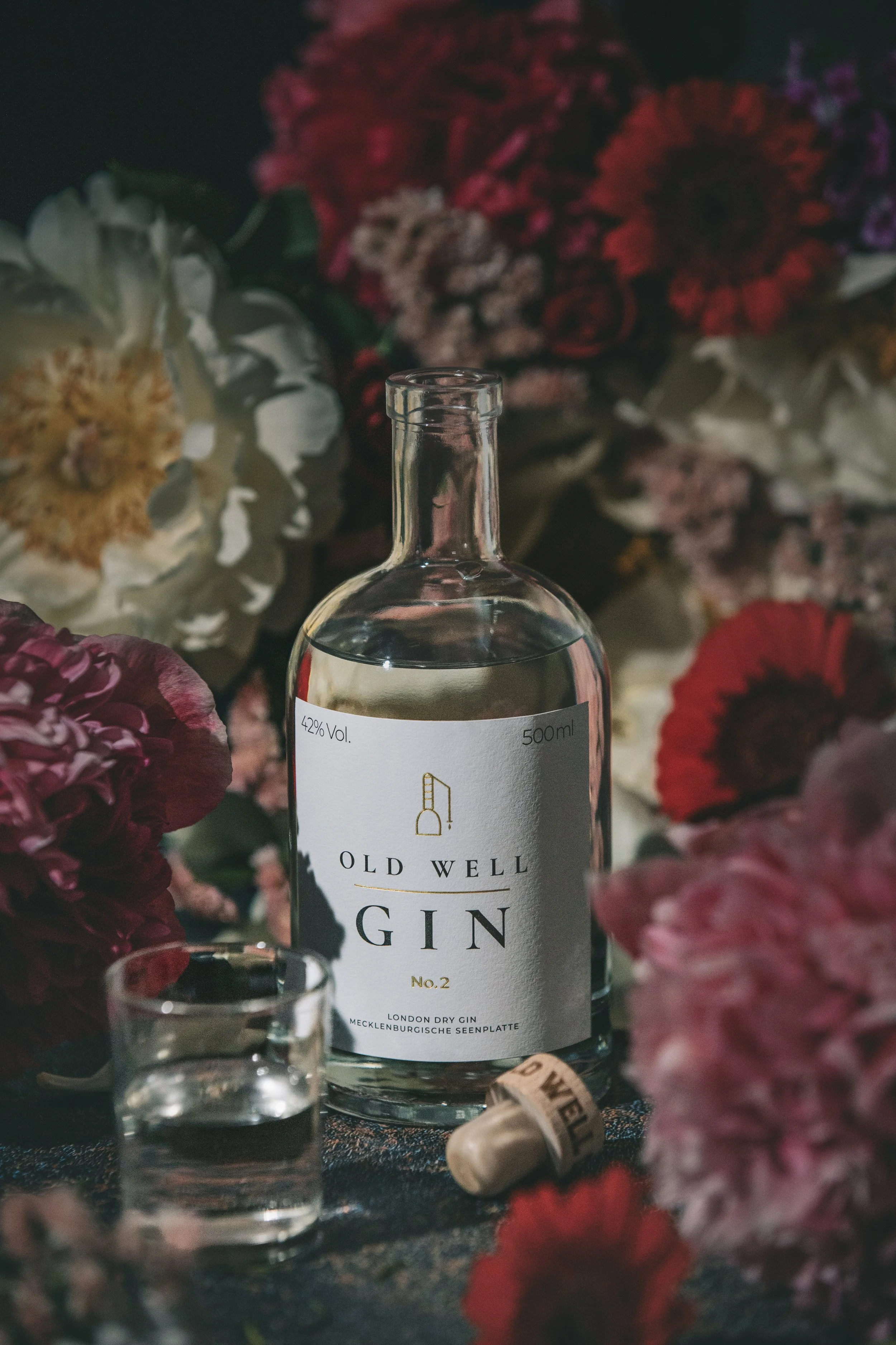 A bottle of Old Well gin surrounded by colorful flowers, with a small glass of gin and a cork in the foreground.