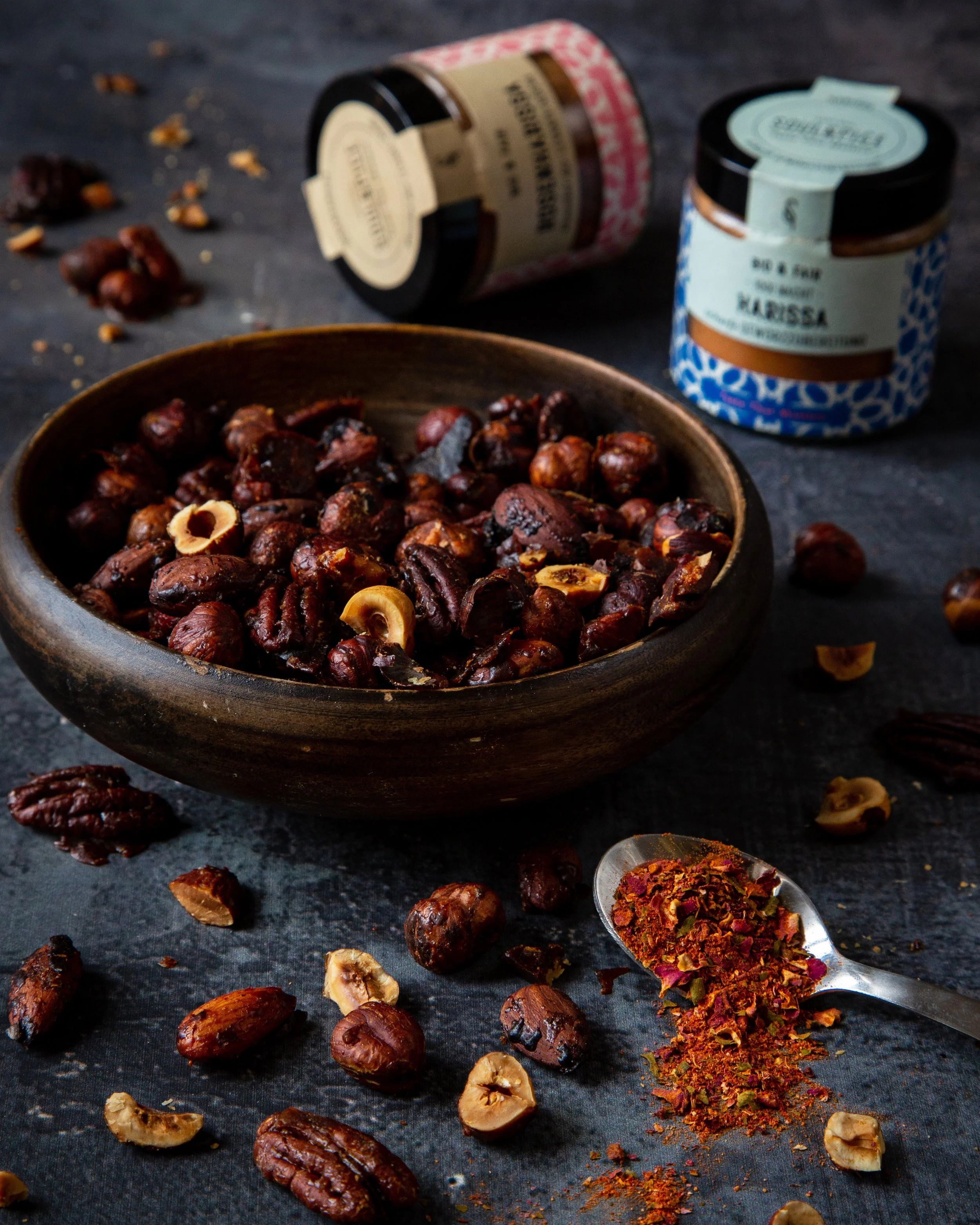 A dark surface with scattered nuts, dried fruit, and spices. In the center, a wooden bowl filled with roasted nuts, mainly pecans and hazelnuts. Two spice jars and a spoon with a colorful spice mix are also on the surface.