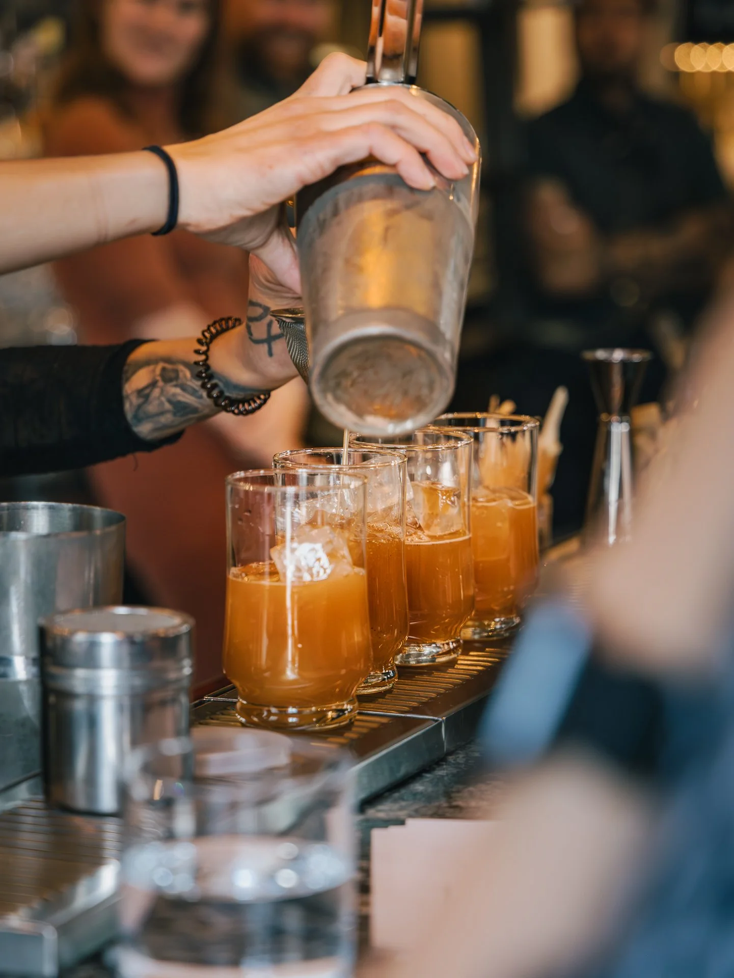 A bartender pours a drink into glasses with ice at a bar, with people in the background.