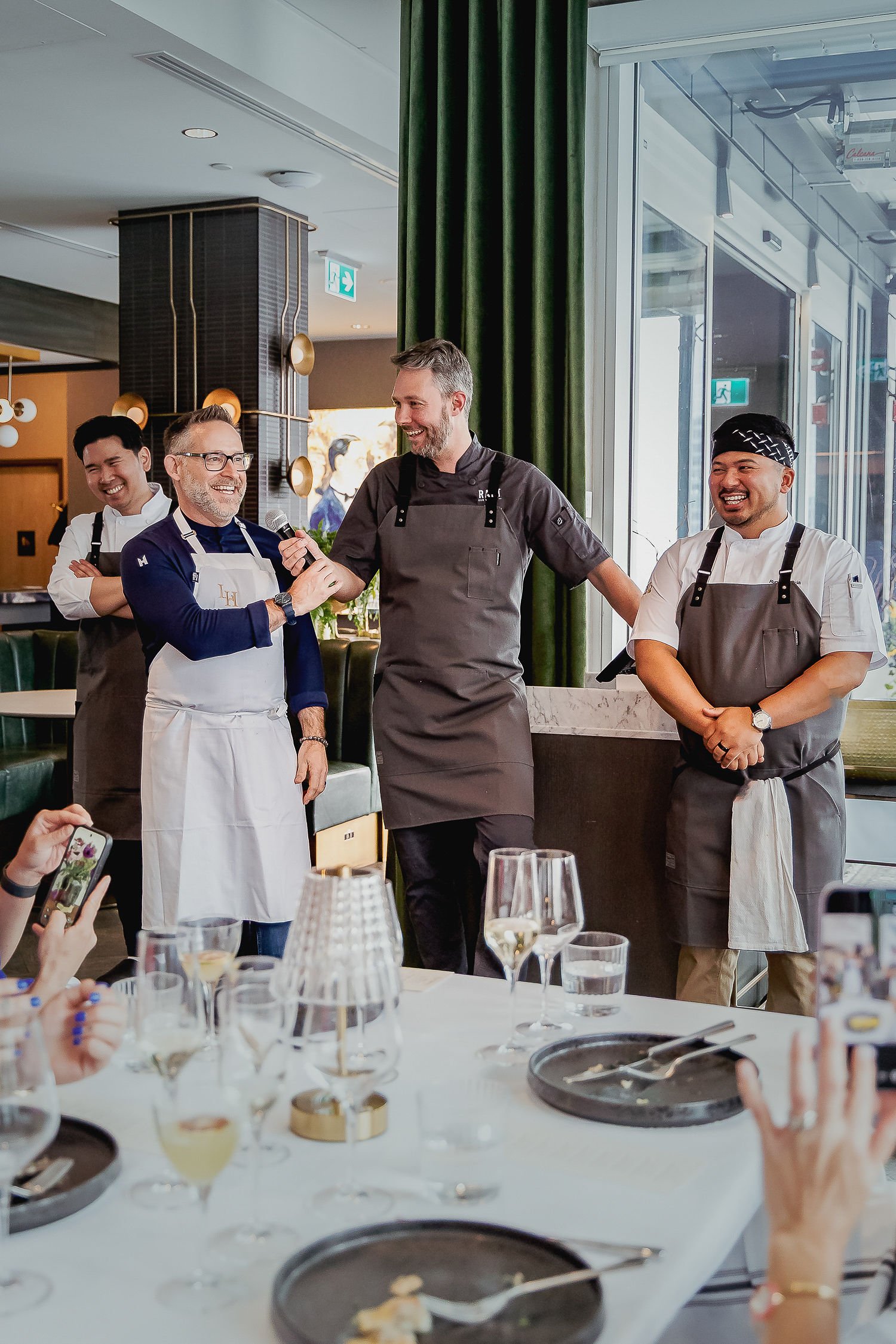 People in a restaurant enjoying a celebration, with a focus on four men standing and smiling, one speaking into a microphone. The table has wine glasses, plates, and utensils, and there are other guests taking photos.