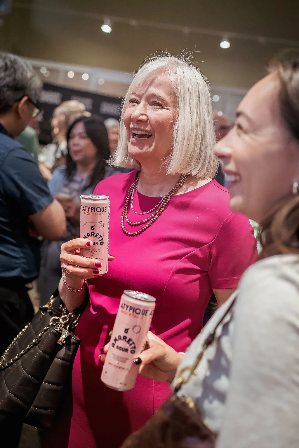 Two women smiling and holding canned drinks at a social event, with a crowd in the background.