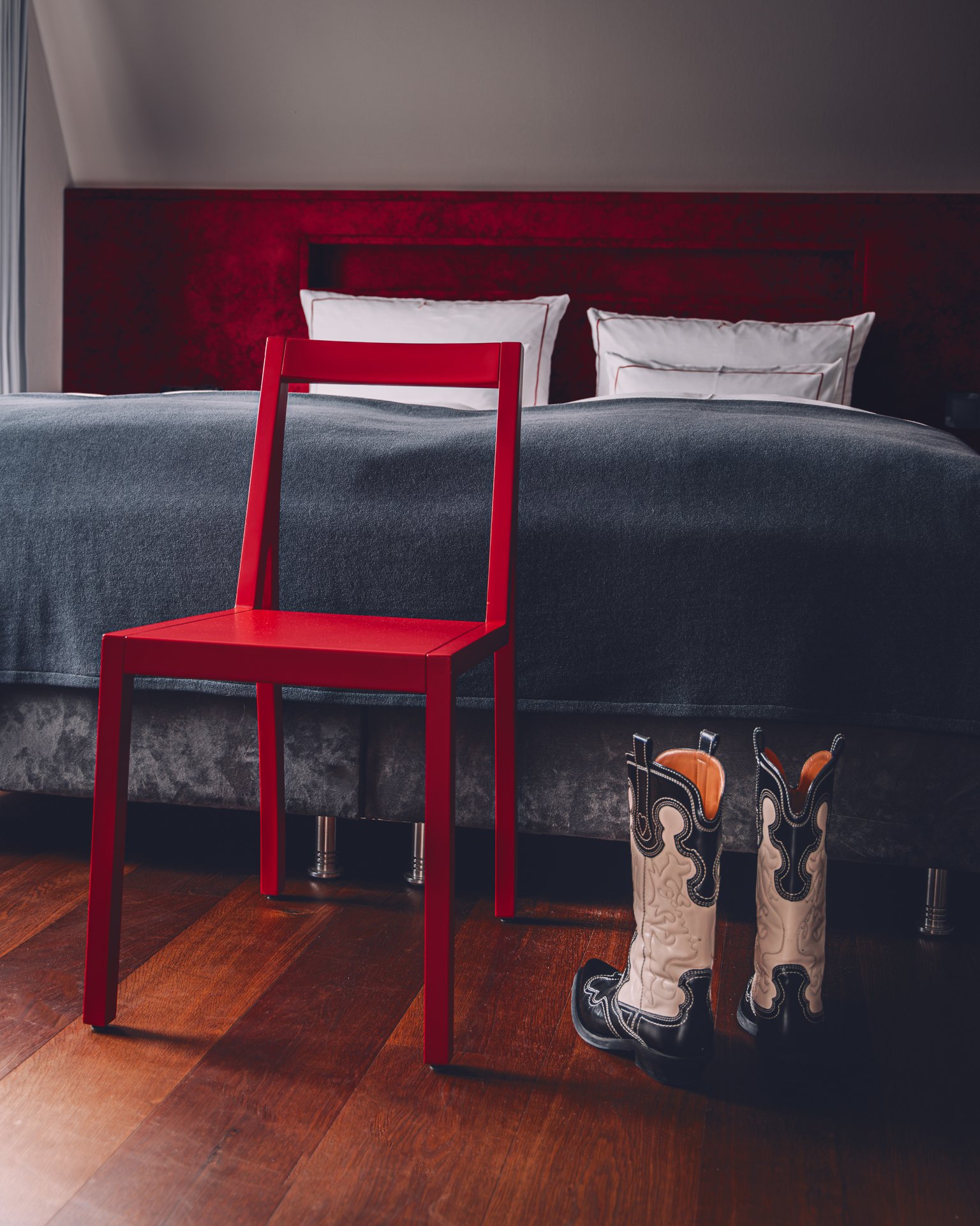 A bedroom scene with a large bed that has a dark gray blanket, four white pillows, and a red headboard. There is a red chair in front of the bed. To the right of the bed, there are cowboy boots with white, black, and tan patterns, standing on a woode