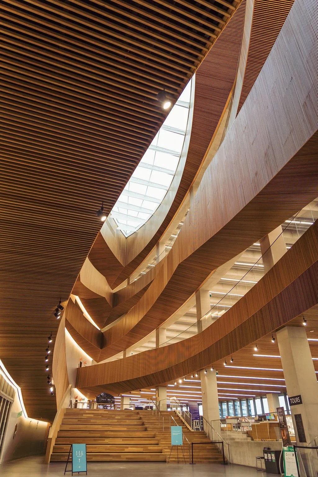 Modern multi-story interior with curved wooden architectural features, large skylight, and staircase, illuminated by ceiling lights.