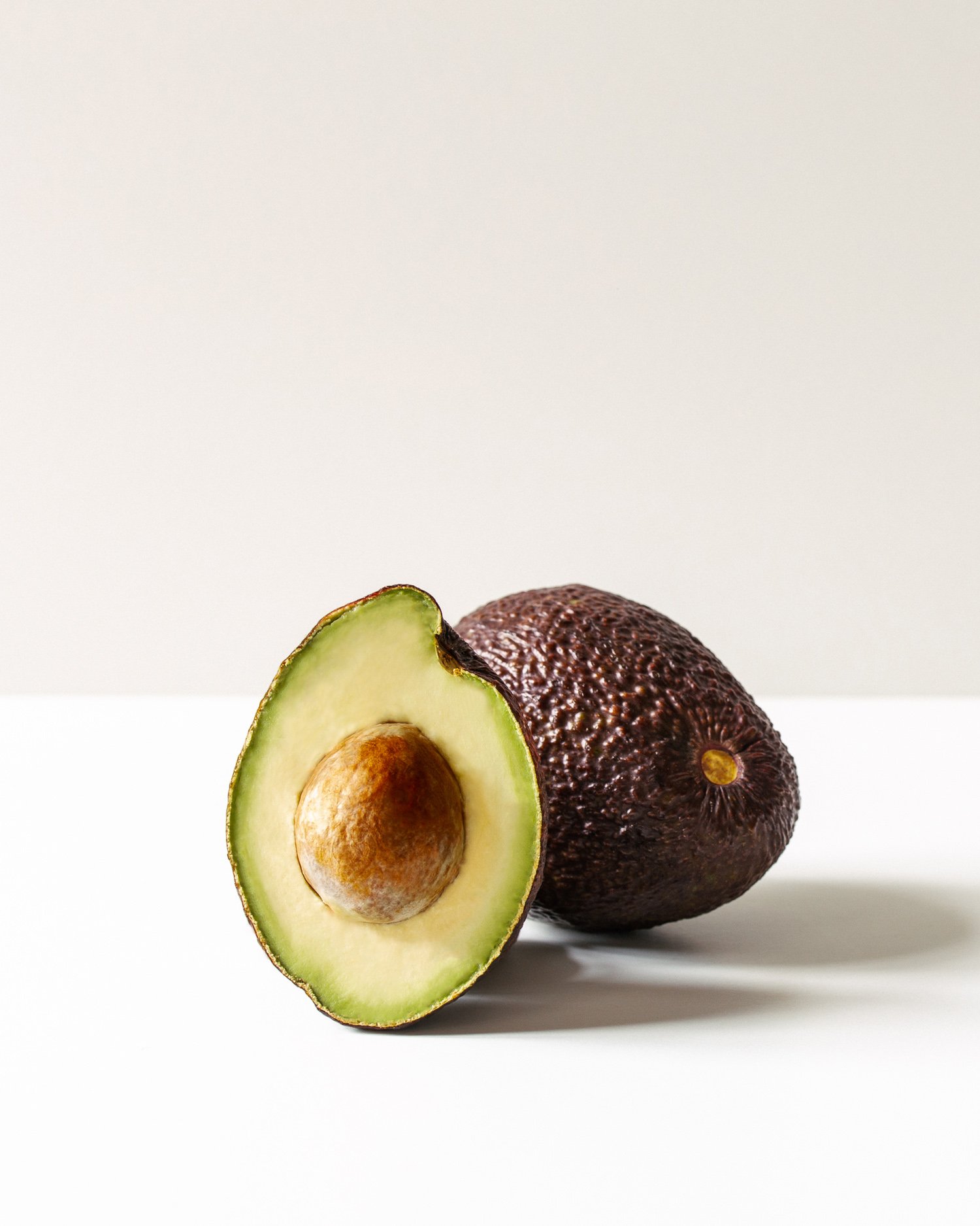 Half avocado with seed in middle and whole avocado behind on white surface.