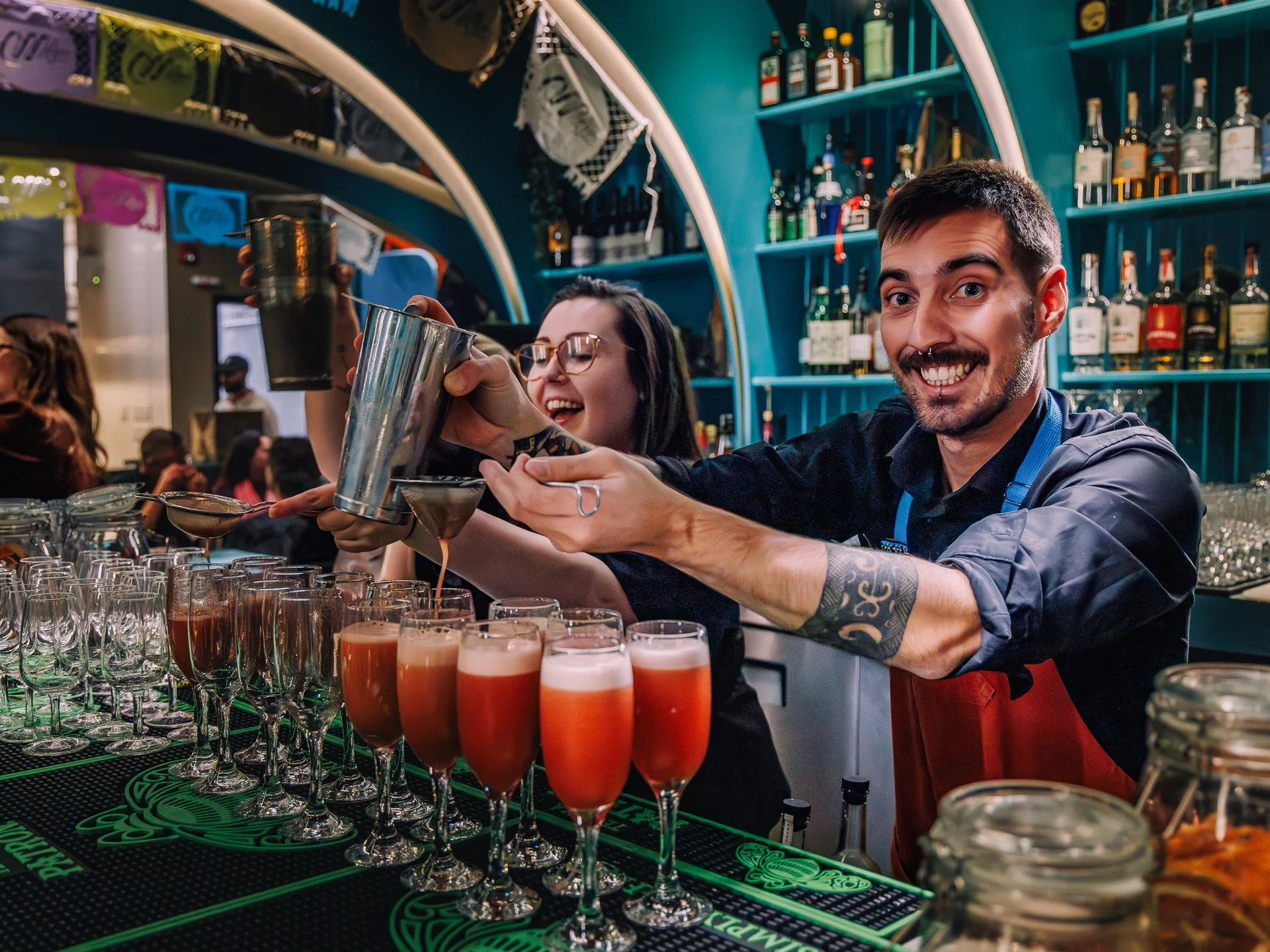 Bartenders preparing colorful layered drinks at a bar with shelves of liquor bottles in the background.