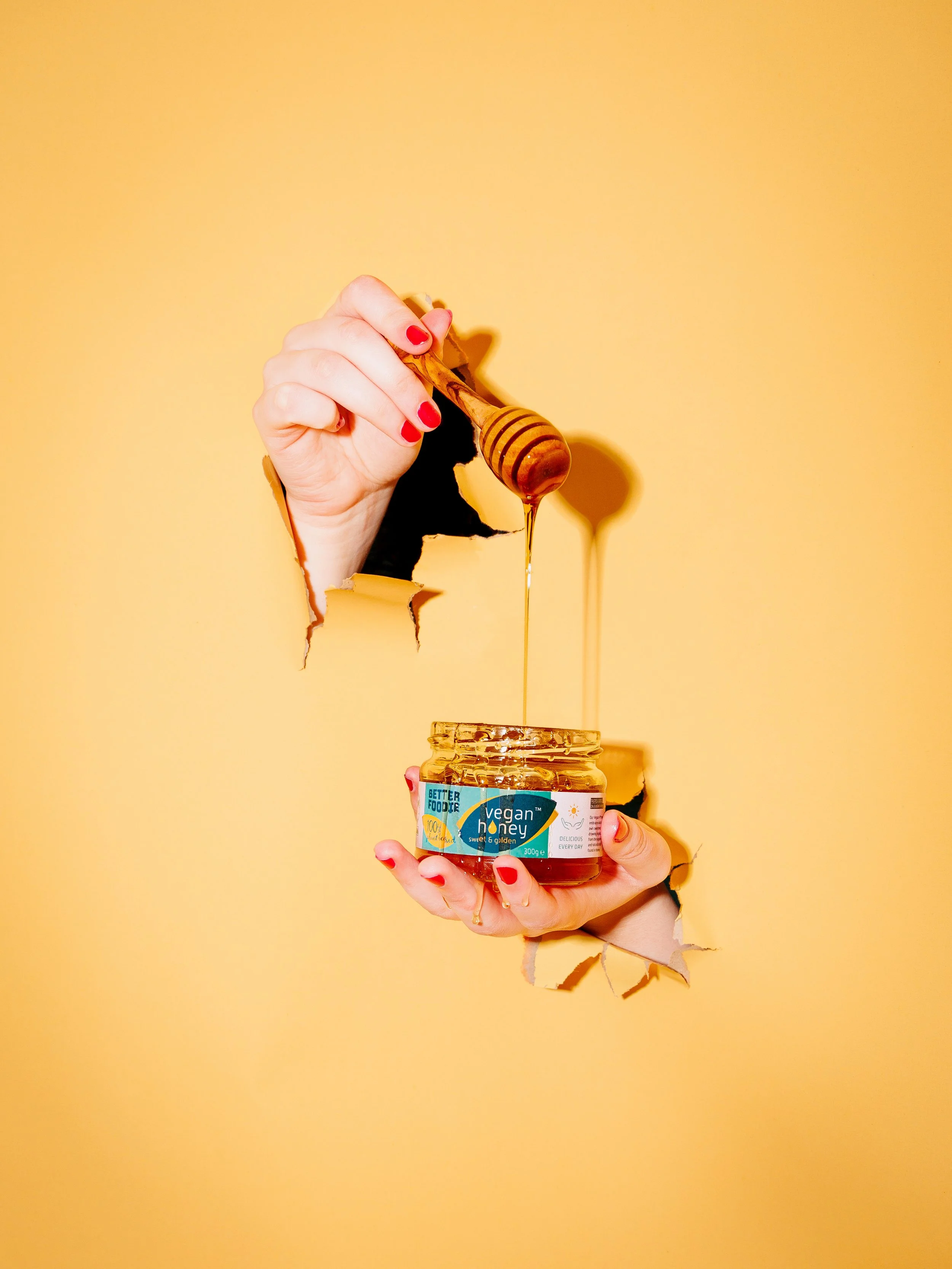 A person holding a jar of vegan honey, with honey dripping from a wooden honey dipper into the jar, against a yellow background, with tear in the wall revealing a hand and part of a face.