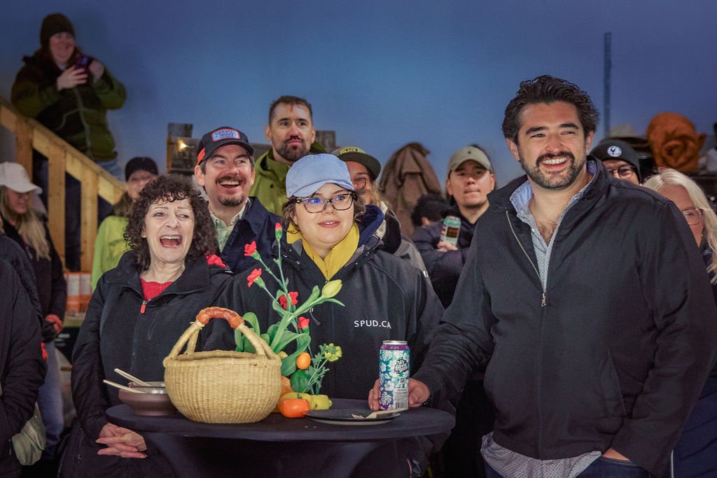 A group of people gathered outdoors at night, smiling and enjoying themselves, with a woman holding a basket and spring-themed decorations on a table.