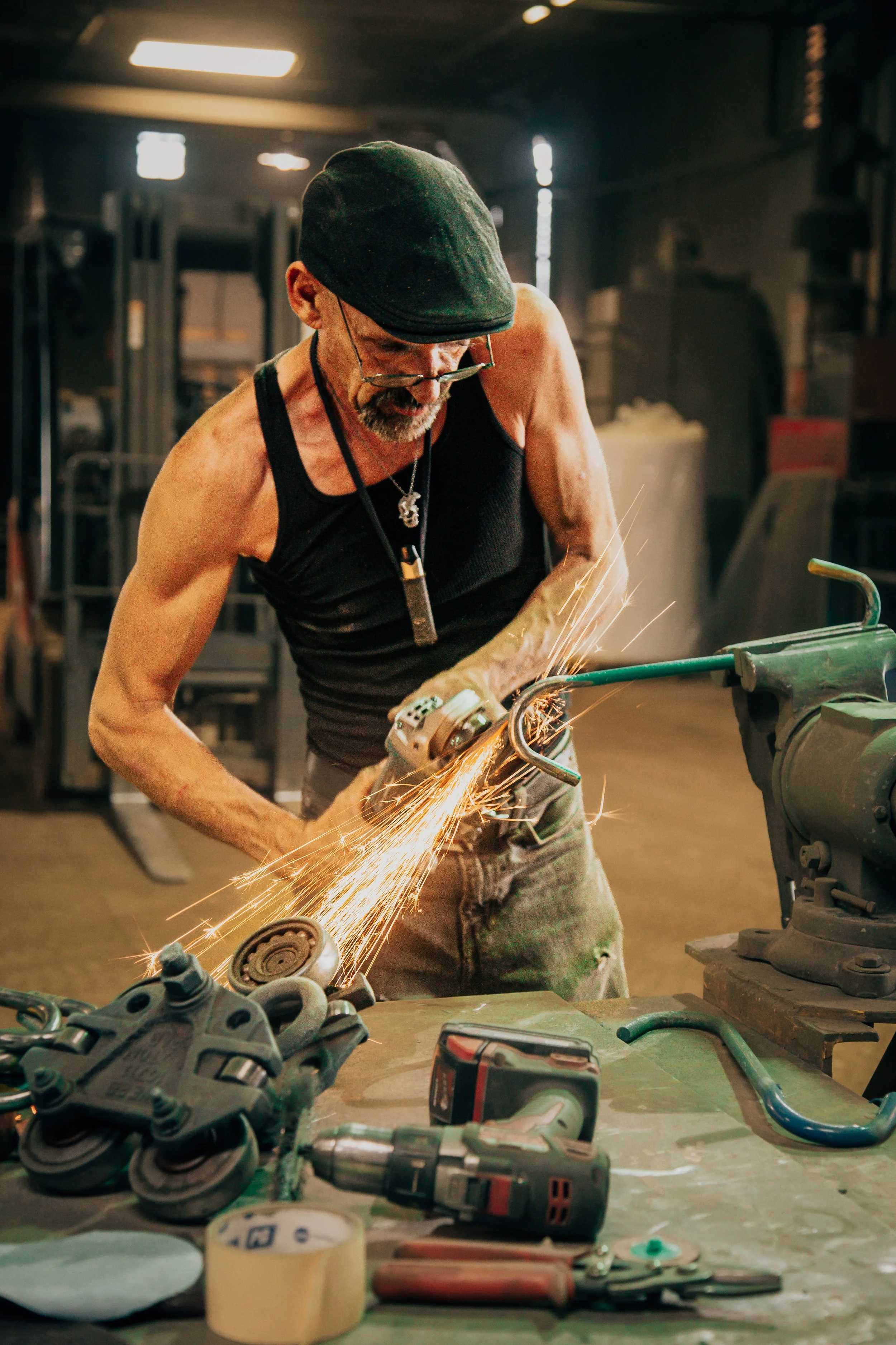 A man wearing a black tank top, glasses, and a cap is working with a grinder, creating sparks in a workshop or garage setting. Various tools and parts are scattered on the table in front of him.