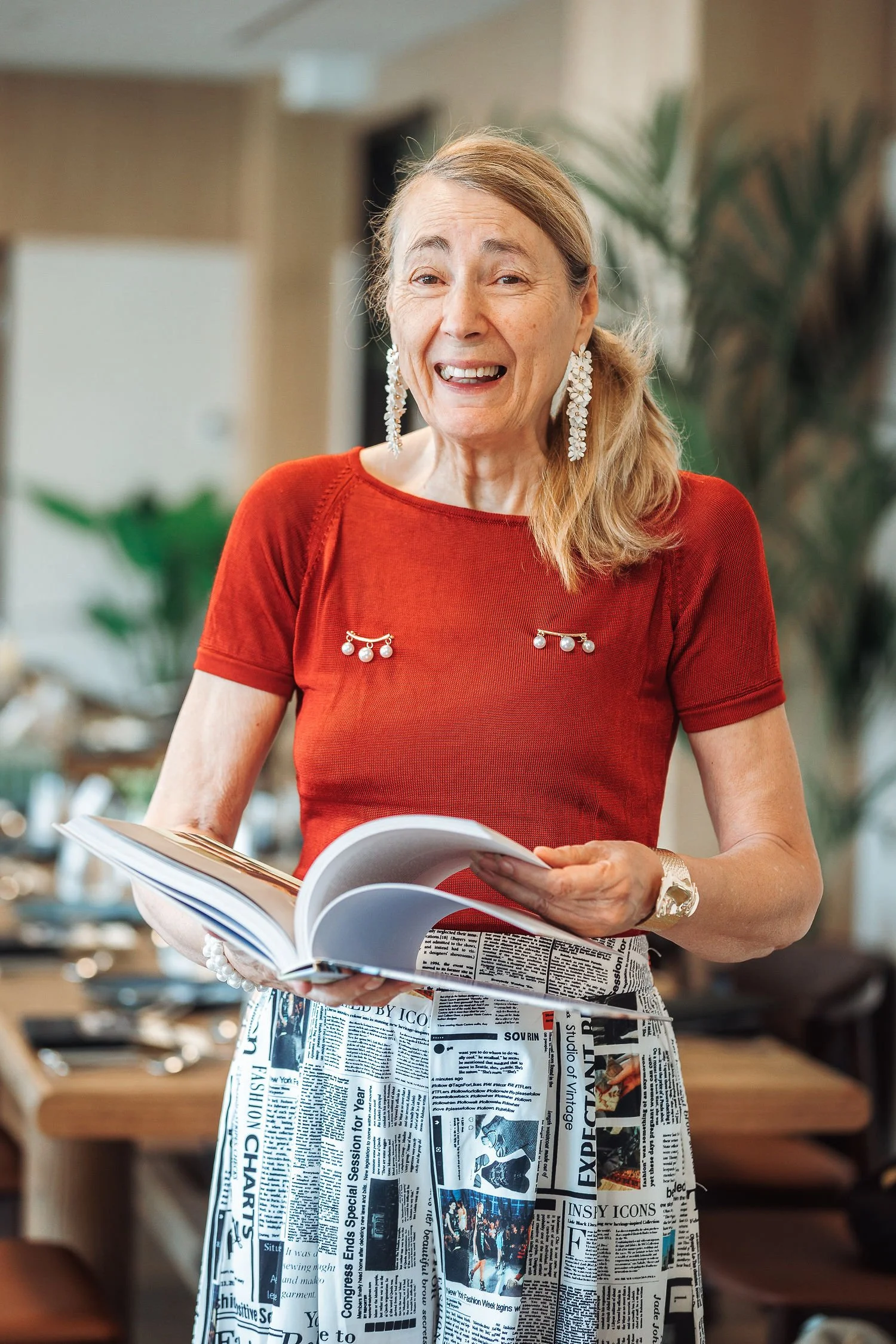 An elderly woman in a red top with pearl earrings is smiling and holding a magazine or newspaper in an indoor setting with plants and furniture in the background.