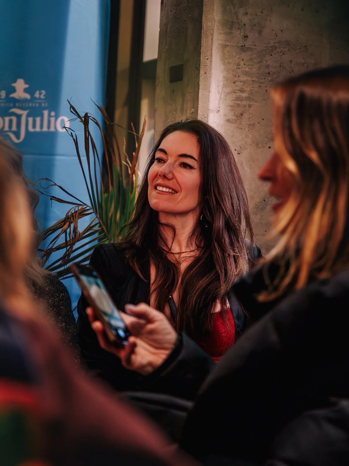A woman with long dark hair smiling and talking to a group of people in an indoor setting with a blue banner and concrete wall