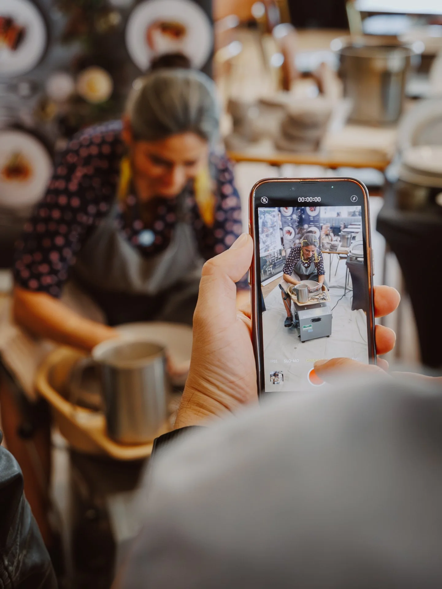 A person taking a photo of a woman working on a pottery wheel in a ceramics studio with a smartphone.