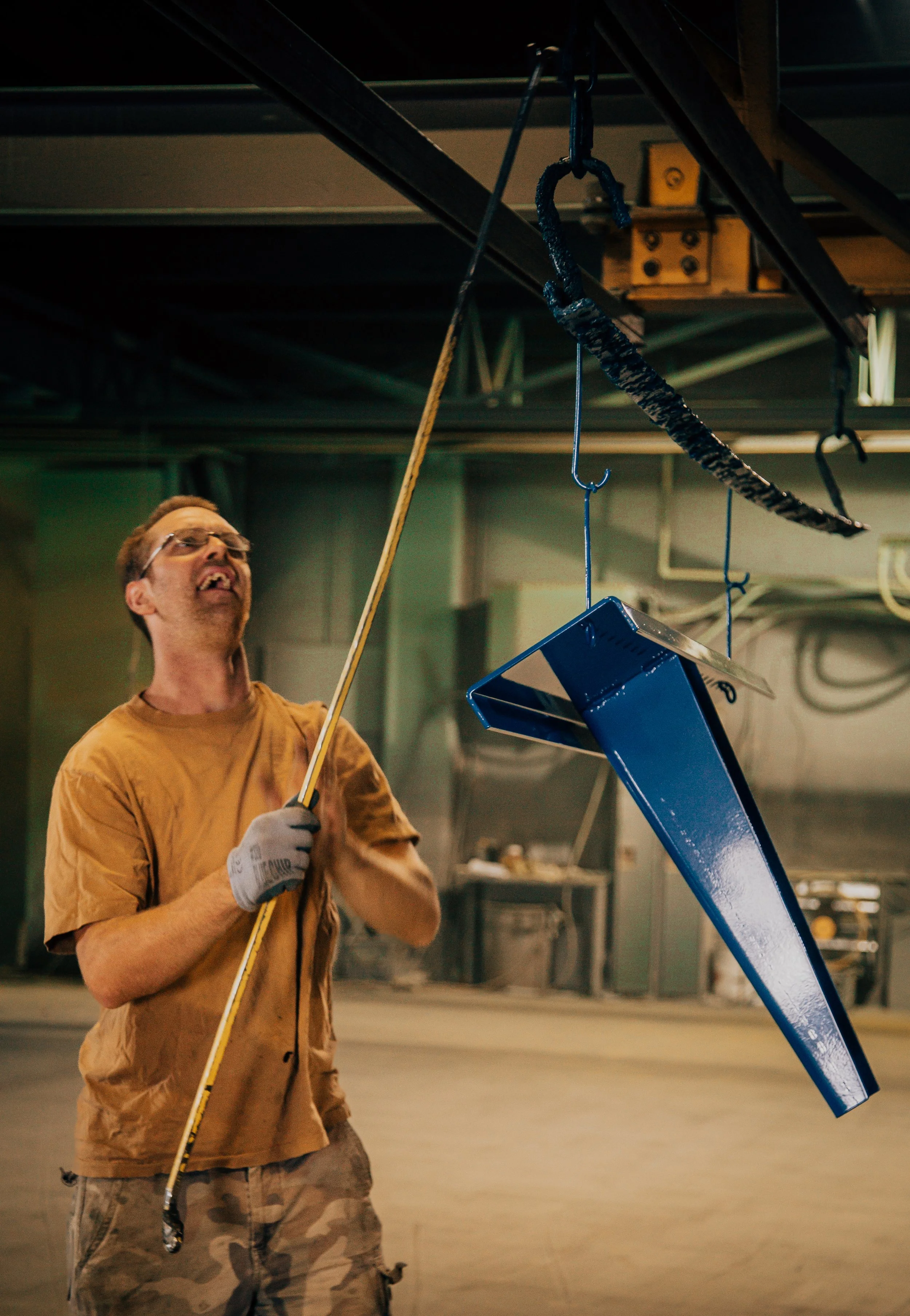 A man wearing glasses and a tan shirt is looking up and smiling while holding a long yellow pole. He is in an industrial or warehouse setting, with a blue metal object hanging from the ceiling using a hook and chains.