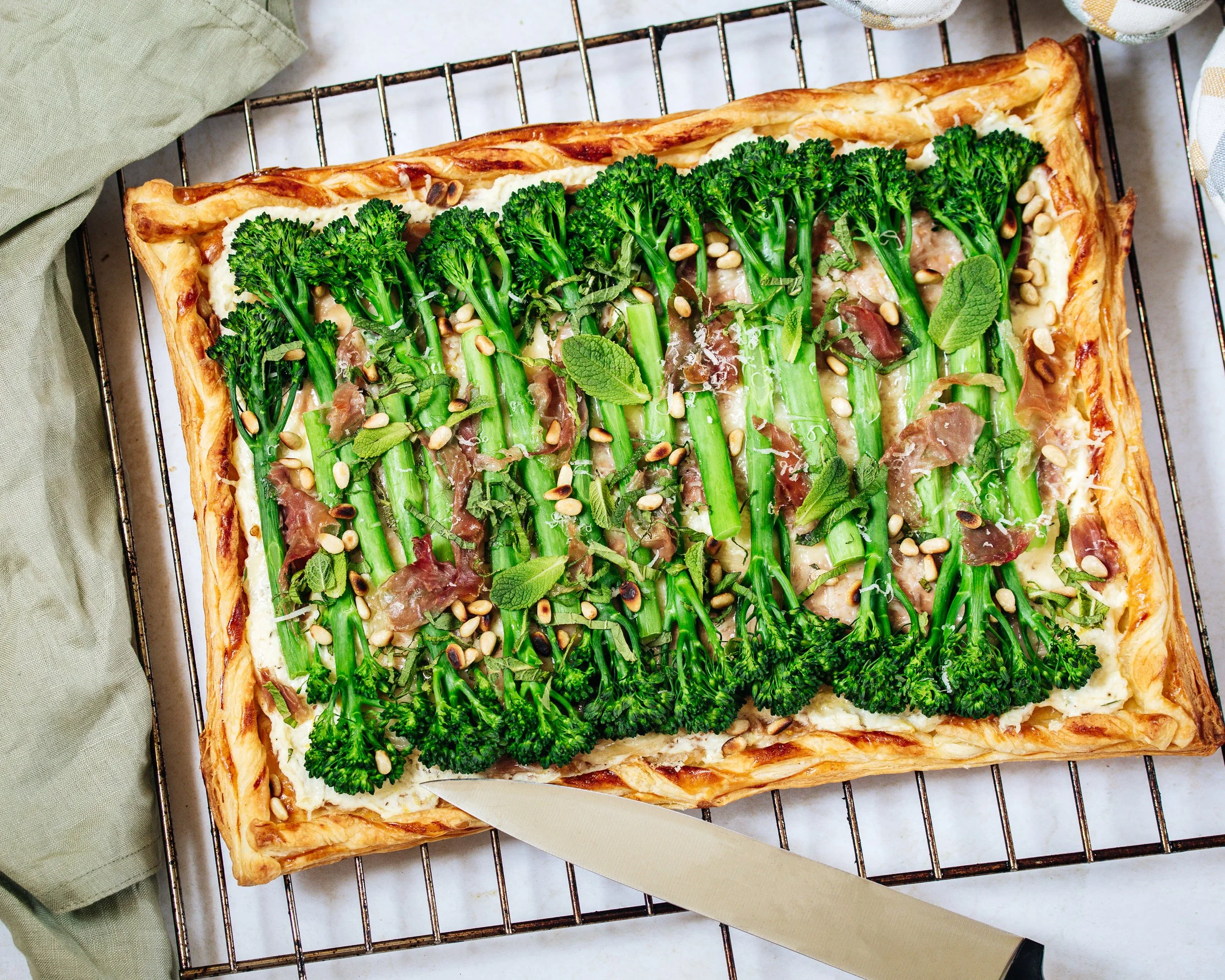 A rectangular vegetable tart topped with broccoli, pine nuts, and herbs on a cooling rack, with a knife and a green cloth nearby.