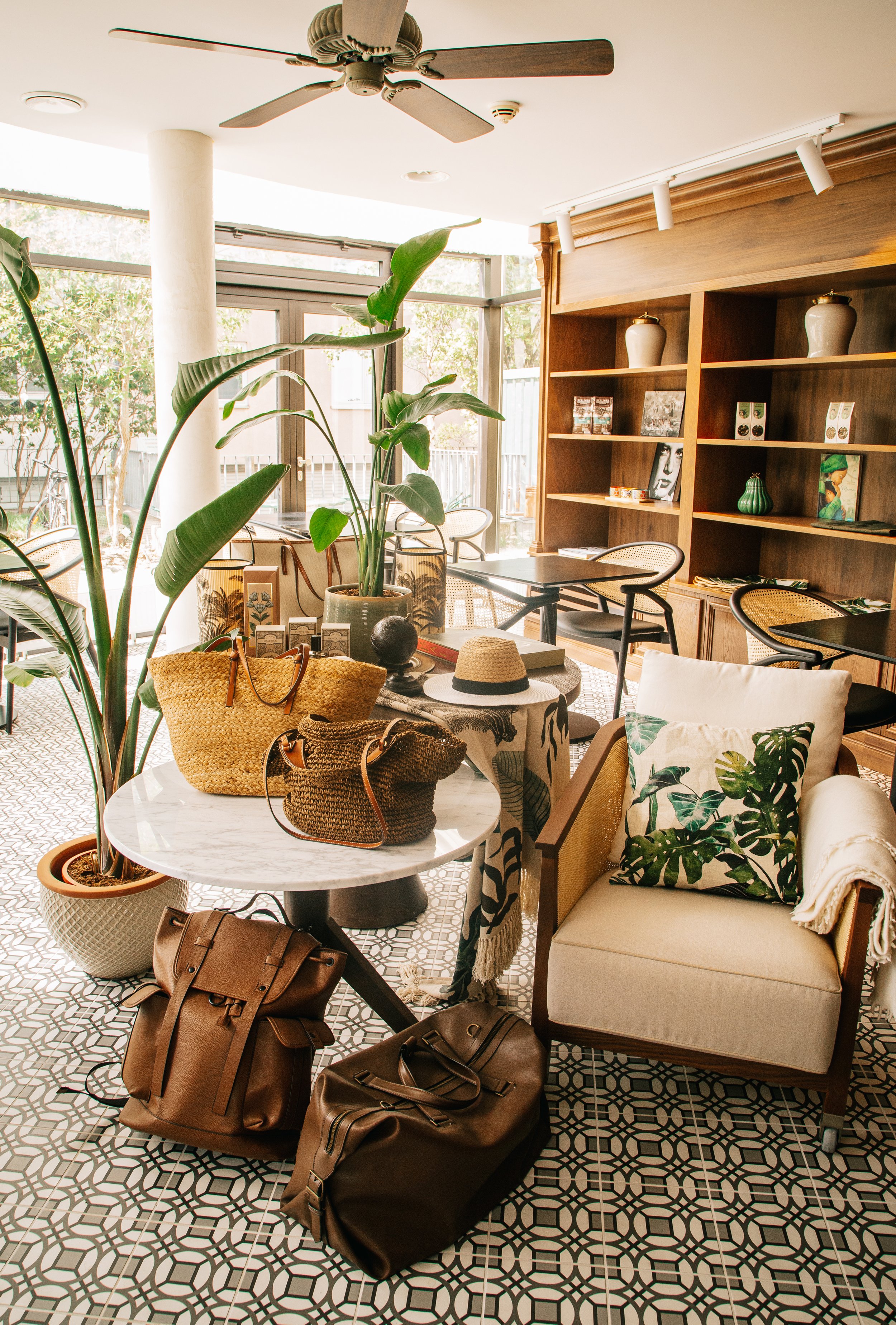Interior of a cozy café with large windows, wooden shelving with decorative vases and books, table with bags and hats, leafy plants, patterned tiled floor, ceiling fan, and chairs with cushions.
