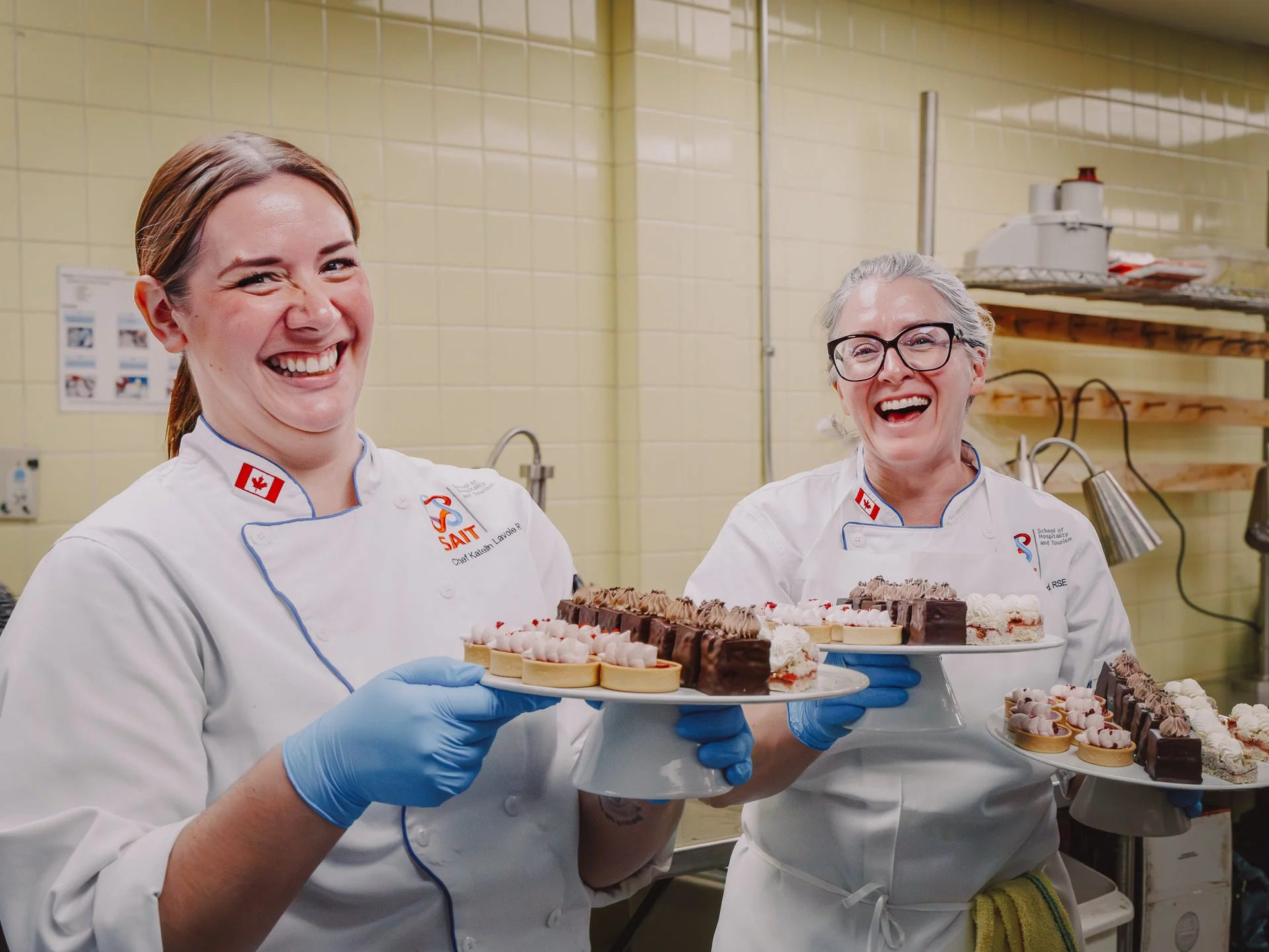 Two cheerful female pastry chefs in chef uniforms with Canadian flag patches, holding trays of assorted decorated cakes and desserts in a kitchen with beige tiled walls and shelves in the background.