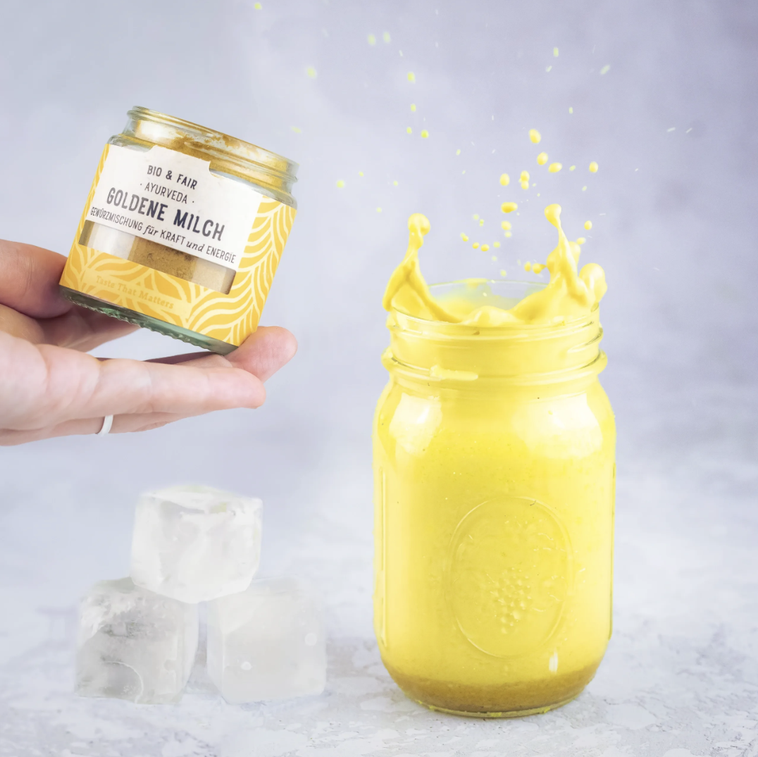 A person's hand holding a small jar of yellow turmeric powder over a mason jar filled with a yellow liquid, with ice cubes on a light gray surface and a plain background.