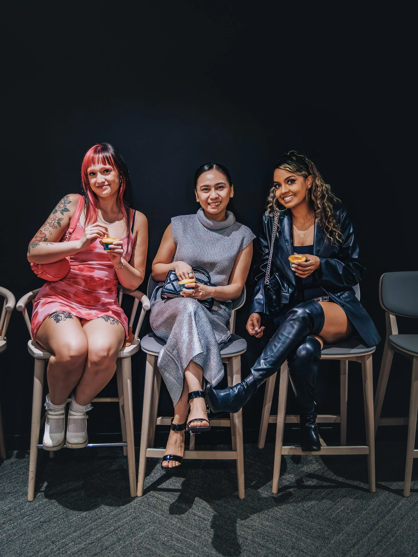 Three women sitting on chairs in front of a black background, smiling and holding small desserts.