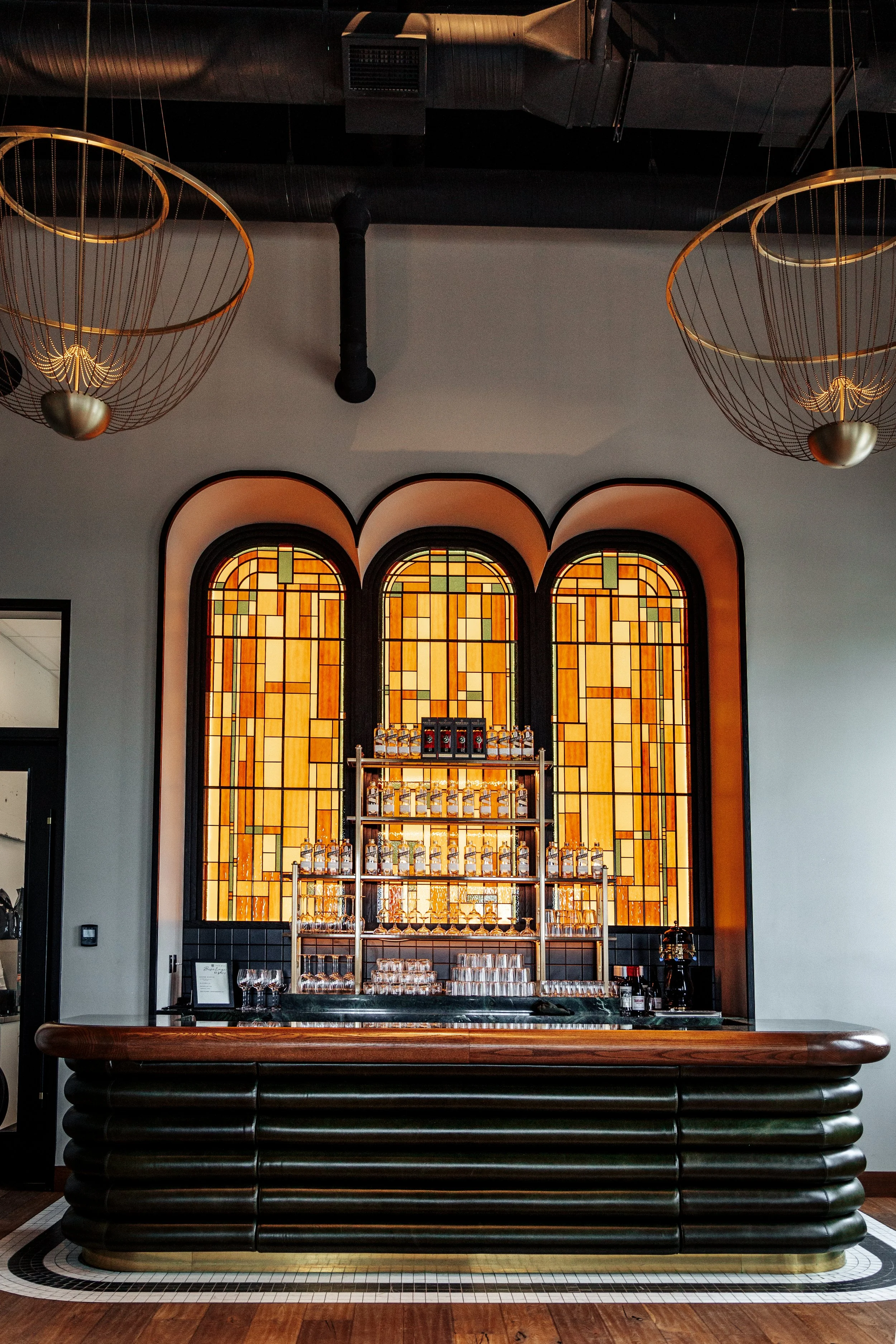 Interior of a bar with stained glass windows behind a black marble countertop, featuring various liquor bottles and glassware, with modern gold wire chandeliers hanging from the ceiling.