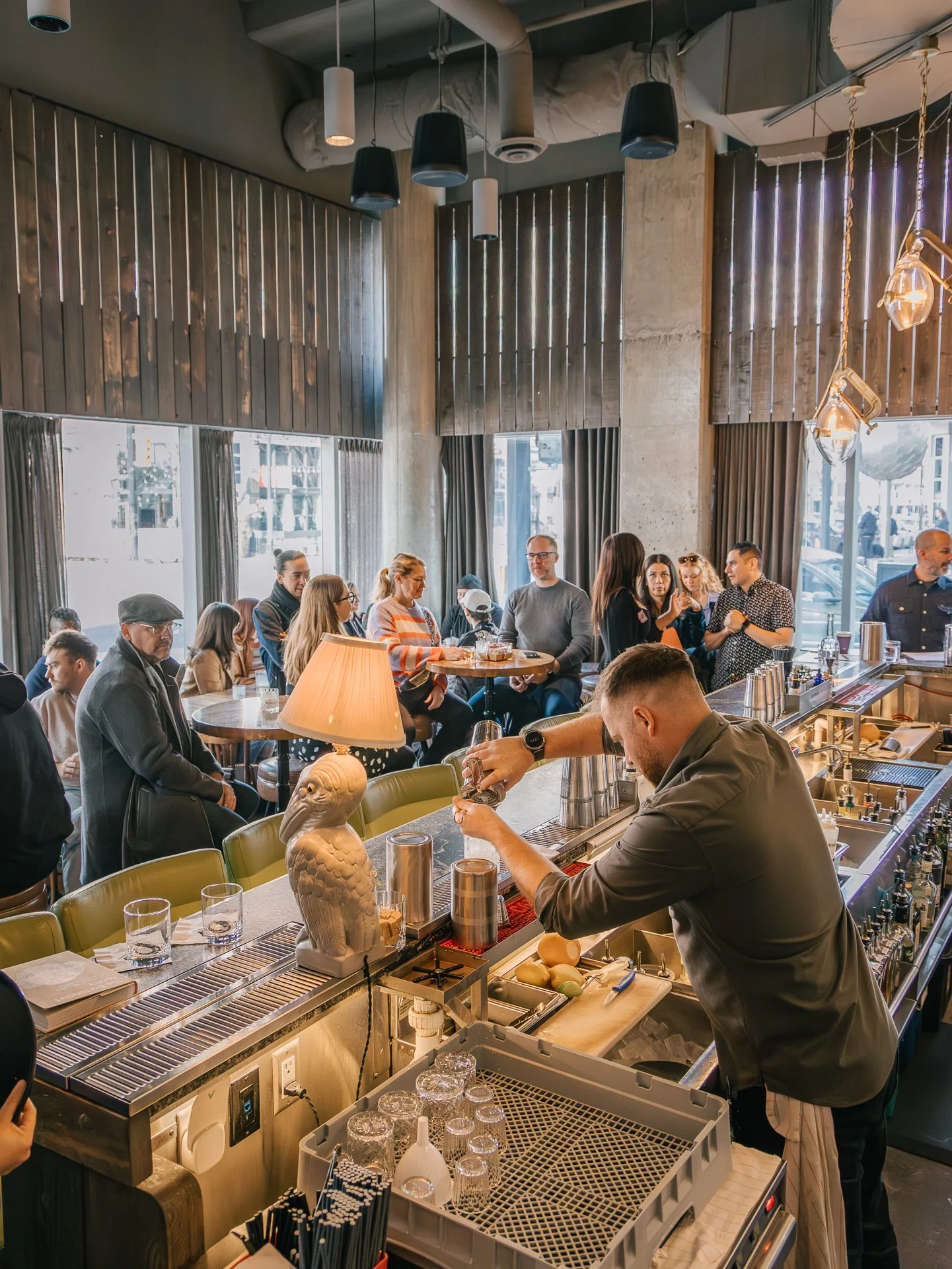 A busy restaurant interior with people sitting at tables and standing near the bar. A bartender is preparing drinks, with various bar tools and decor including a owl sculpture and hanging lights. Large windows let in natural light, and the ceiling ha