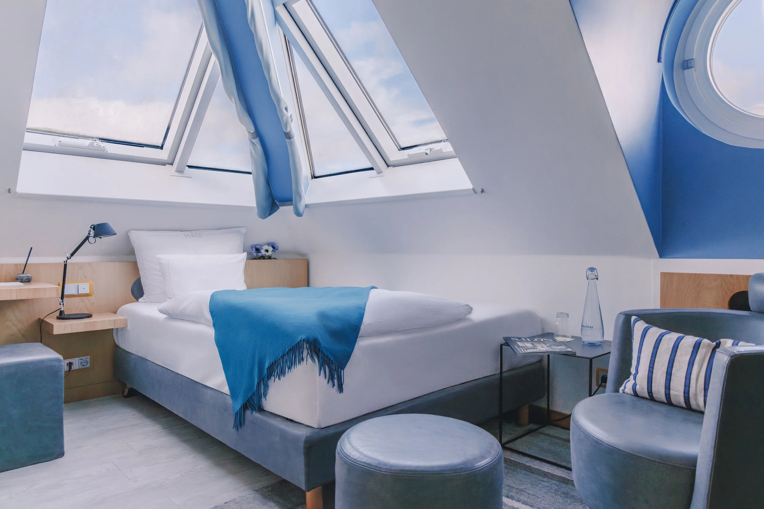 A bedroom with a bed featuring white linens and a blue throw blanket, situated beneath a skylight window with curtains. The room has modern decor, a nightstand with a lamp, a glass of water, a magazine, and a blue footstool.