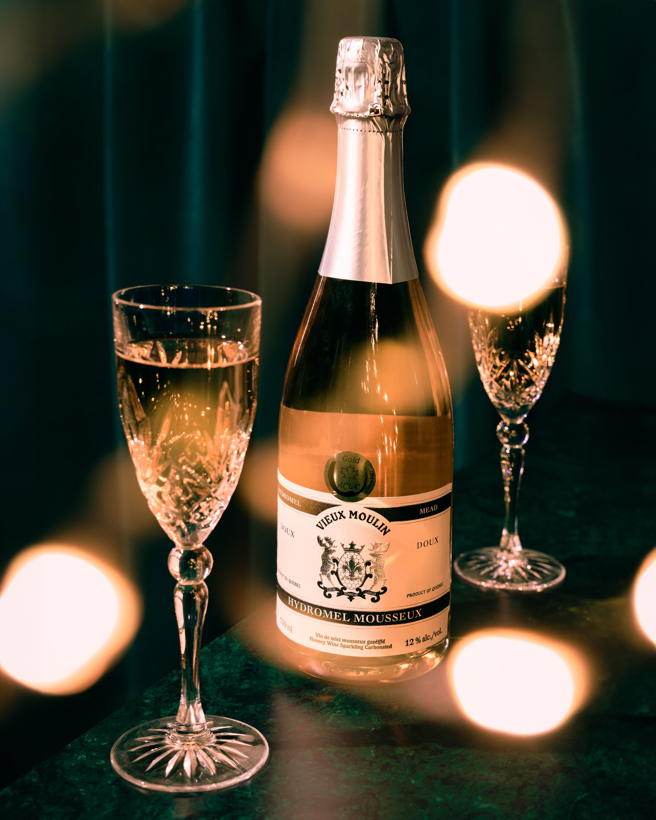 A bottle of sparkling wine with two crystal champagne glasses on a dark surface, illuminated by soft, warm light with blurred round bokeh lights in the background.