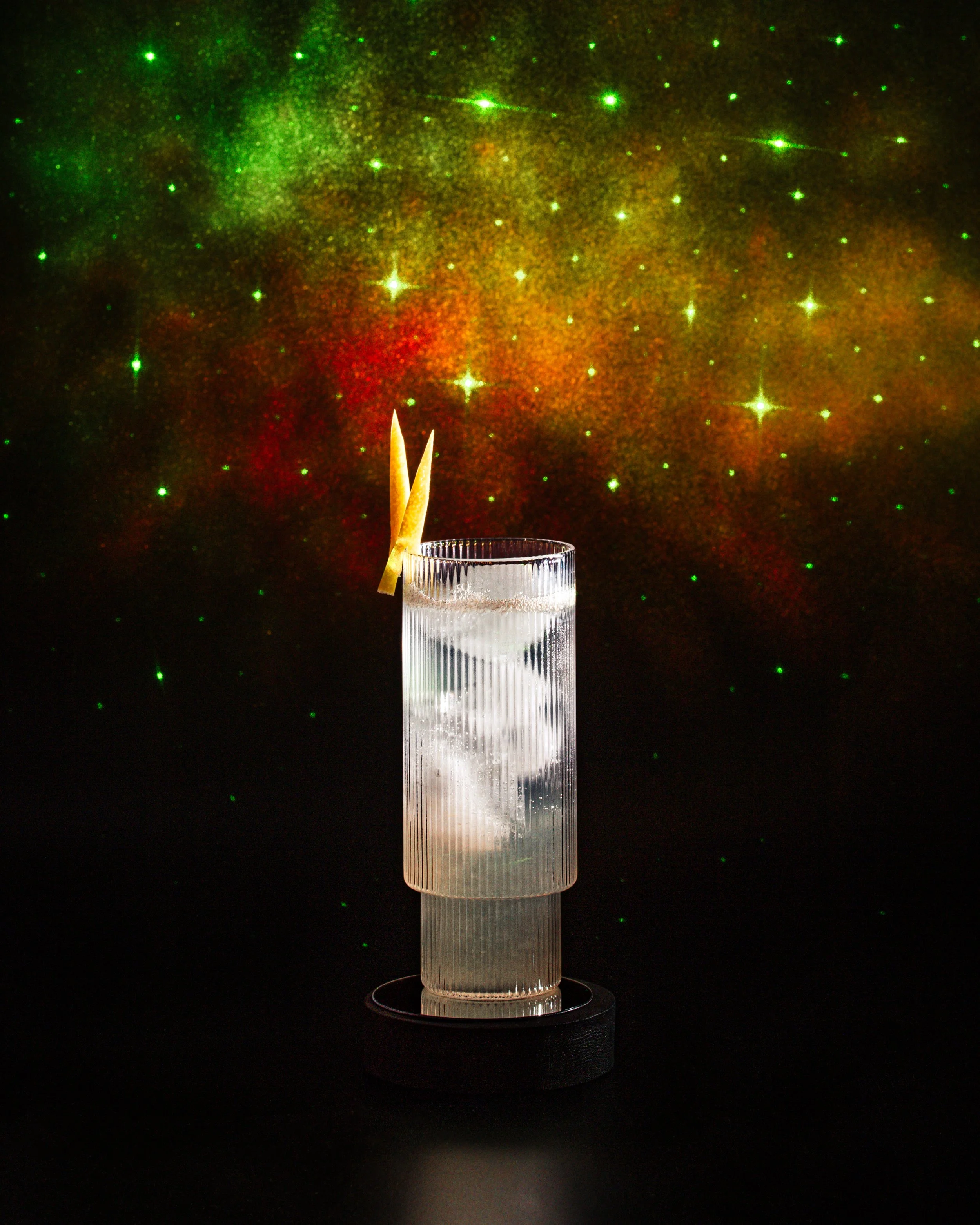 A champagne flute is filled with clear liquid and garnished with a decorative yellow stick. The background features a colorful galaxy pattern with green, red, and yellow stars and cosmic clouds.
